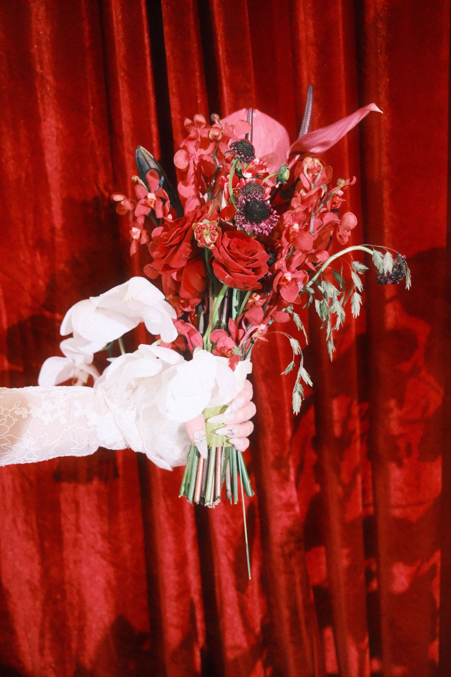 A gloved hand holds a red floral bouquet with roses against a rich red velvet curtain backdrop.