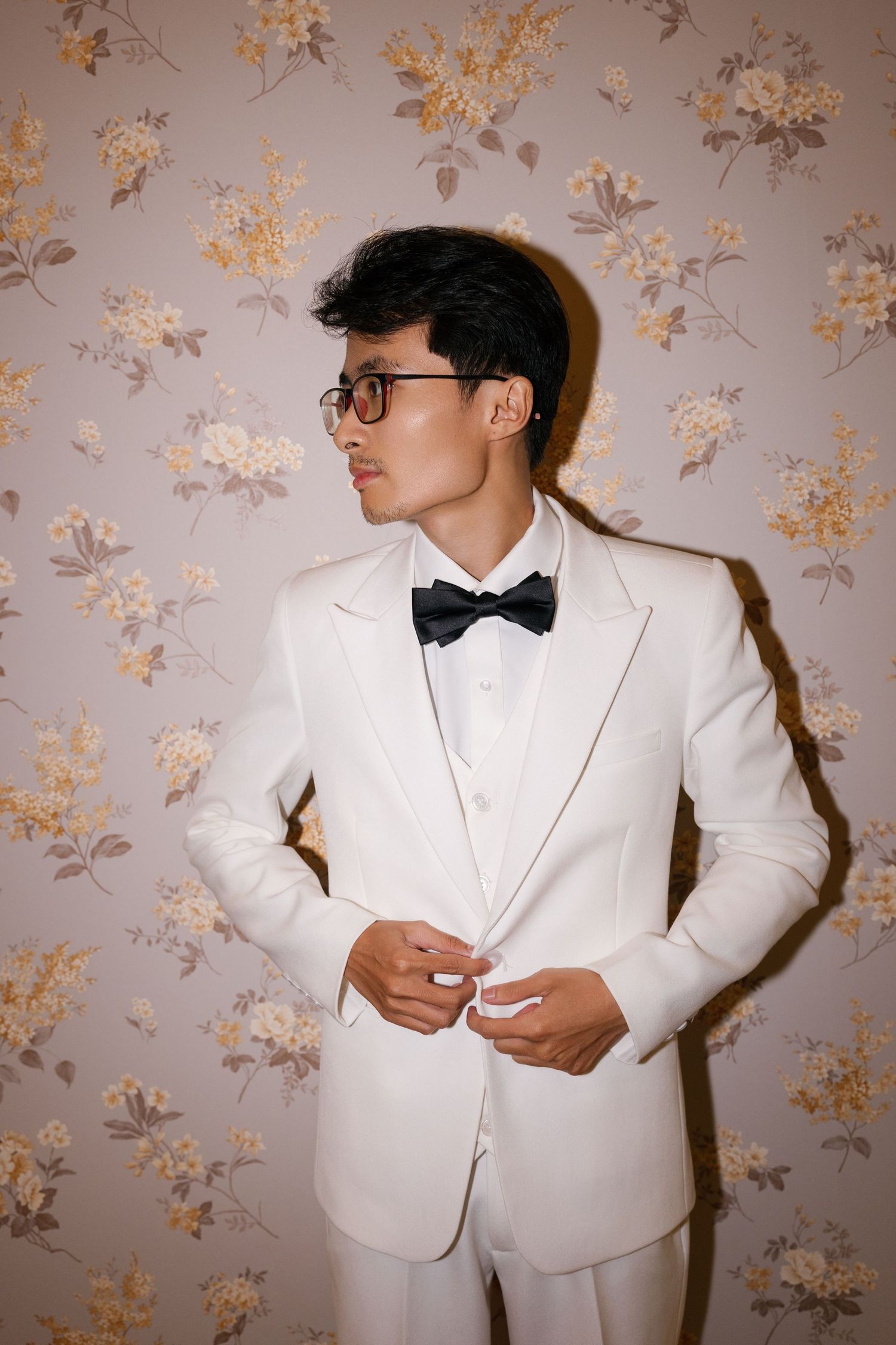 Young man in white tuxedo with black bow tie buttoning jacket against floral wallpaper background.