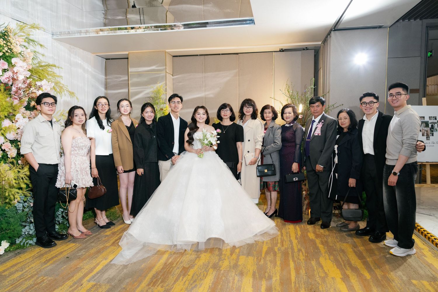 A bride in a white ballgown poses with family and friends at an elegant wedding reception venue decorated with floral arrangements.