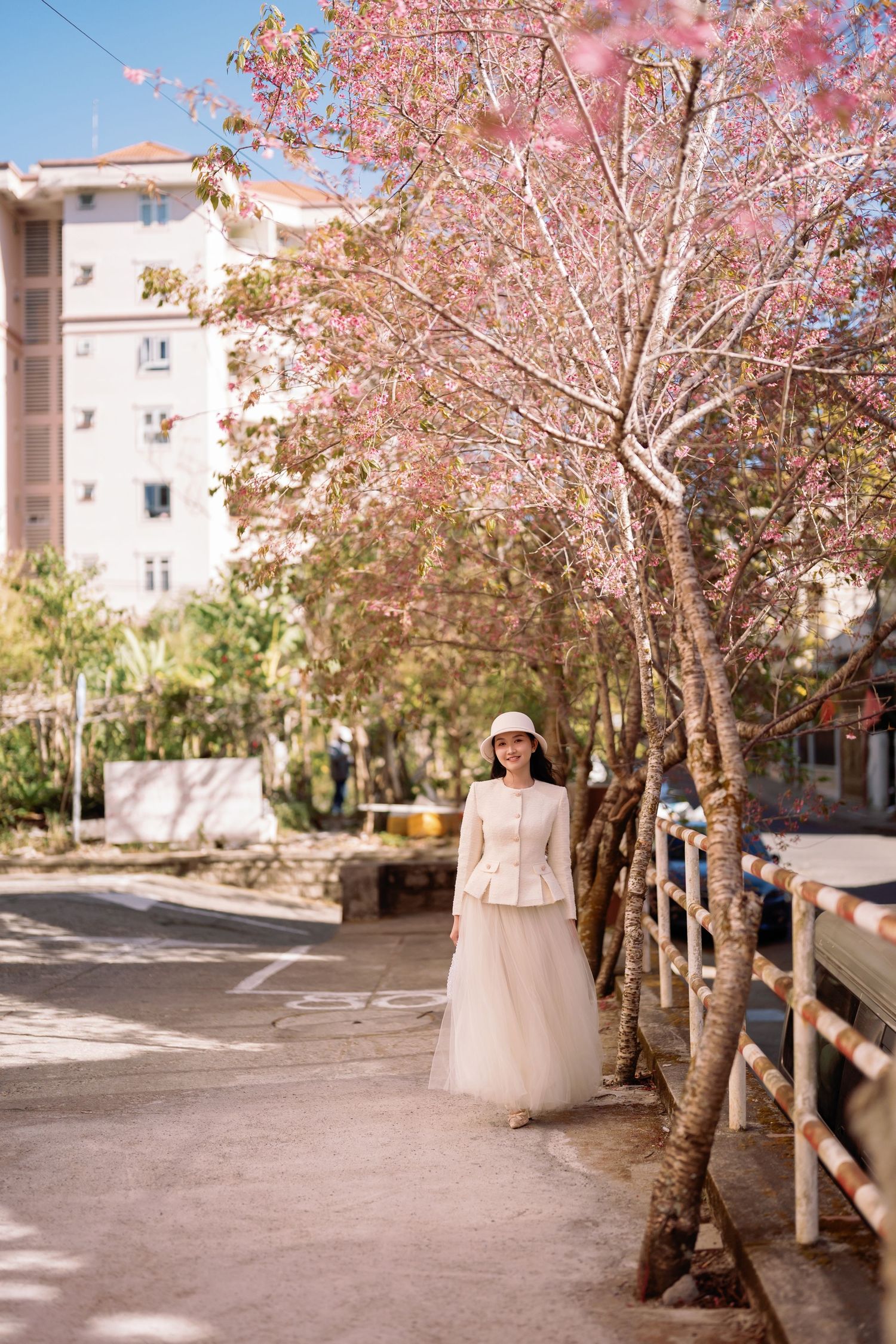 Woman in white outfit and hat stands under pink cherry blossom trees on a sunny urban street.