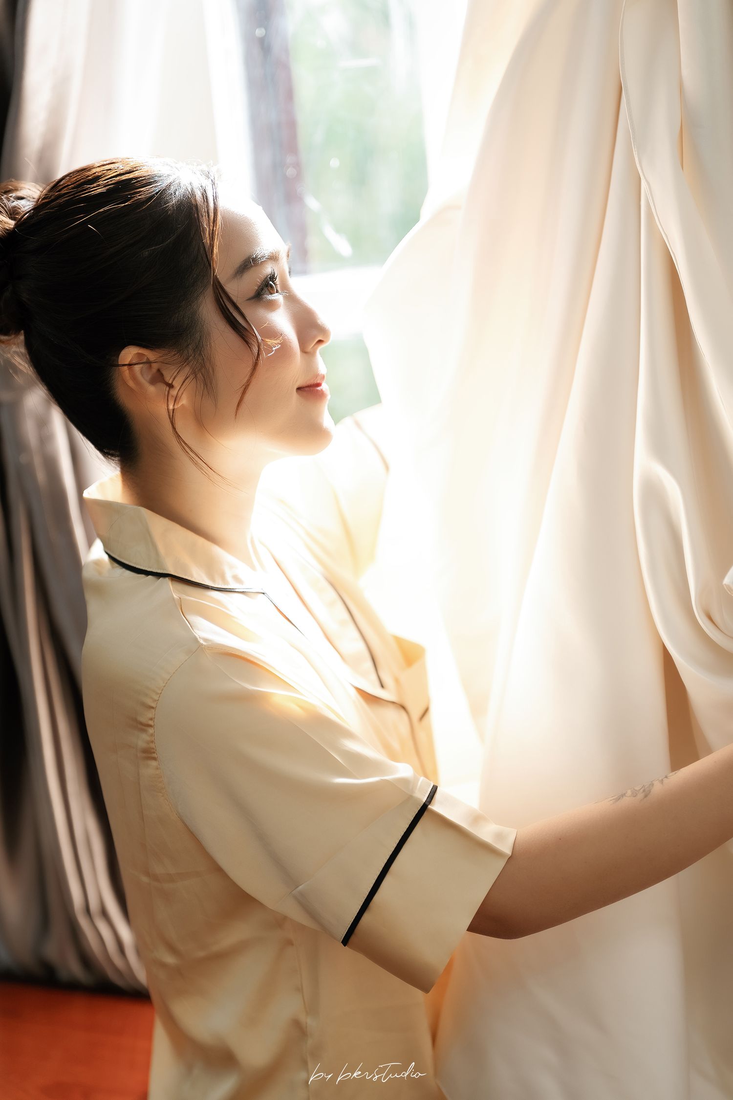 A young woman in a yellow shirt gazes upward while holding white curtains near a sunlit window.