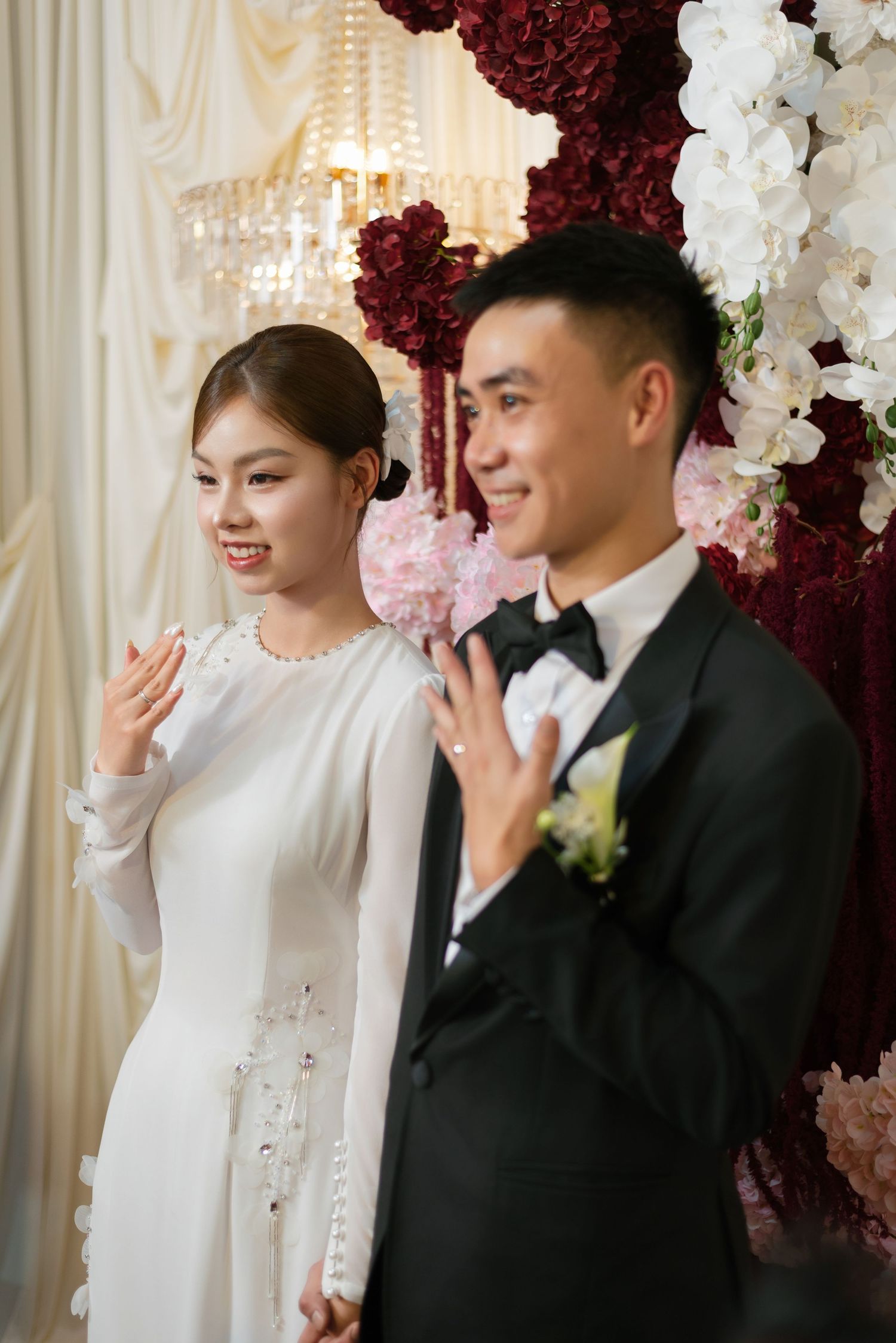 A bride in white ao dai and groom in black tuxedo wave and smile before a floral arch of red and white flowers.