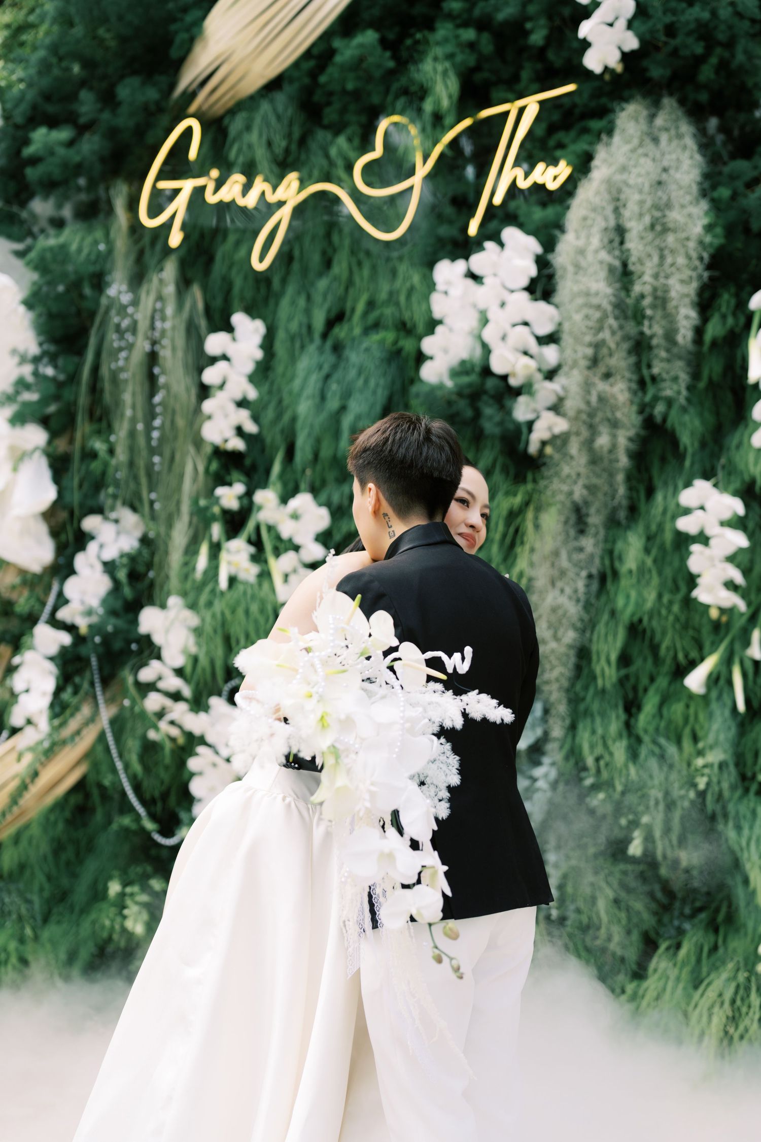 Bride and groom embracing before green floral wall with Giang and Thao neon wedding sign.