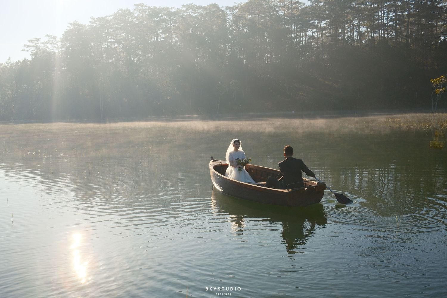 Bride and groom in a wooden rowboat drifting on a serene lake surrounded by pine trees in morning light.