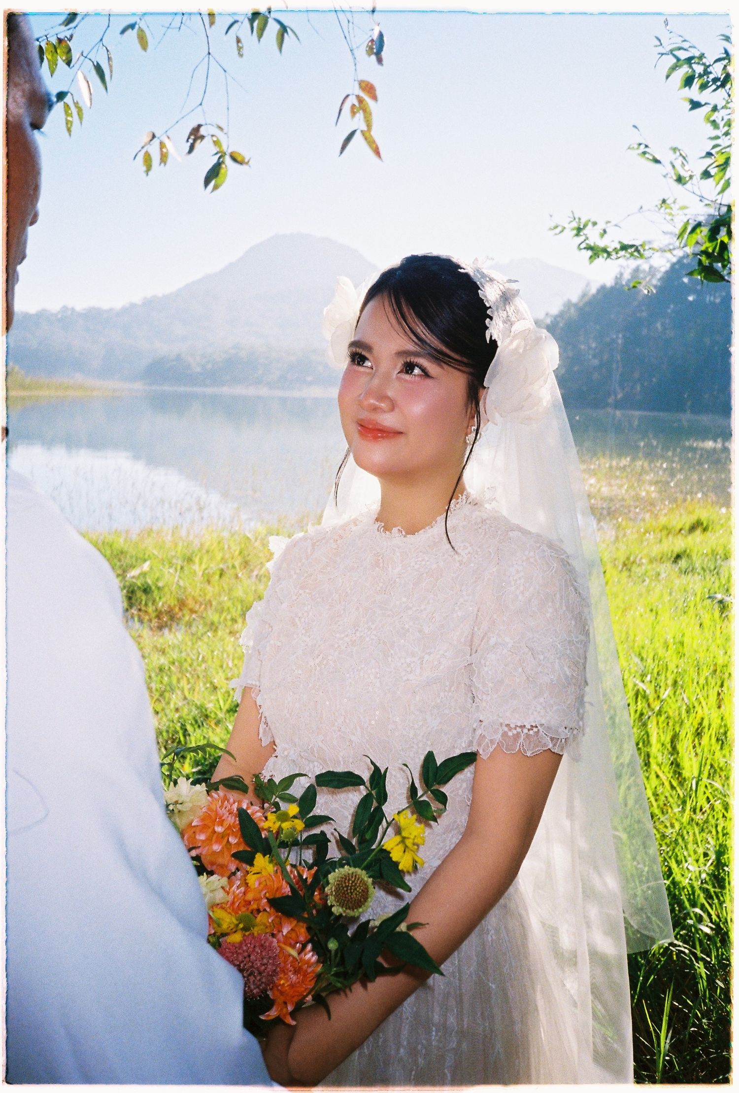 Smiling bride in lace dress holds bouquet with mountain lake scenic backdrop on sunny wedding day.