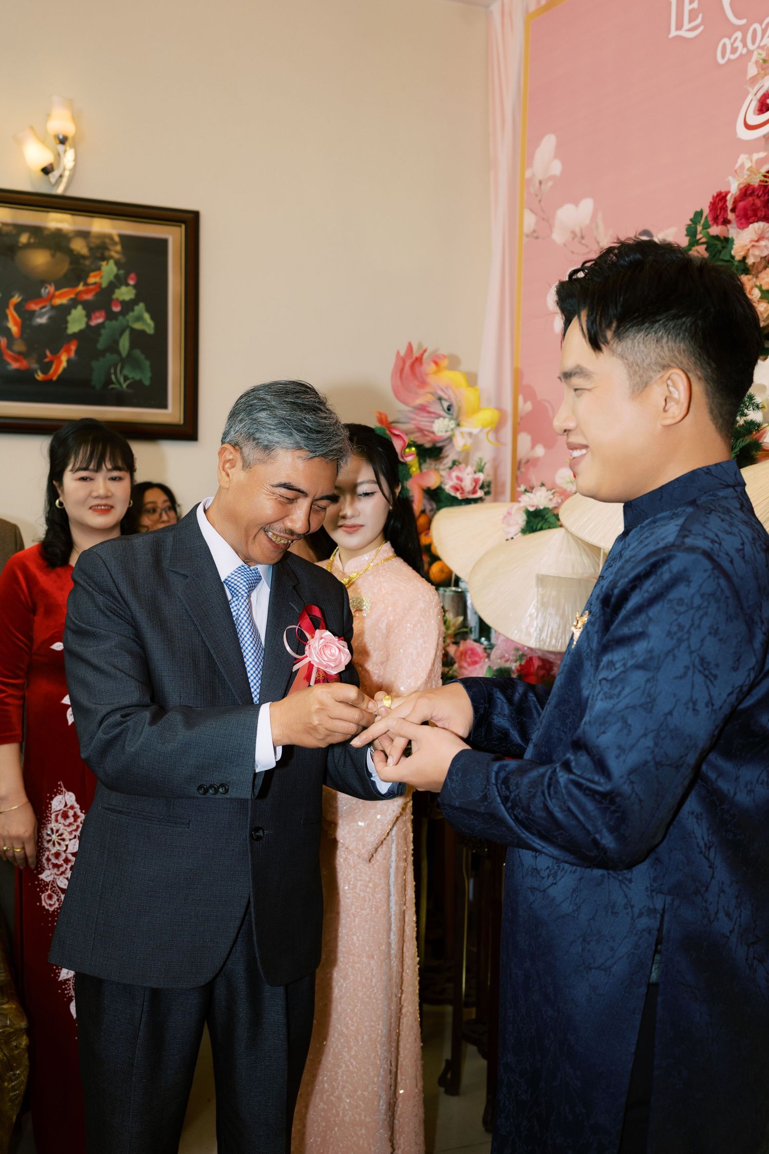 Older man in suit placing ring on young groom in blue ao dai at traditional Vietnamese wedding ceremony.