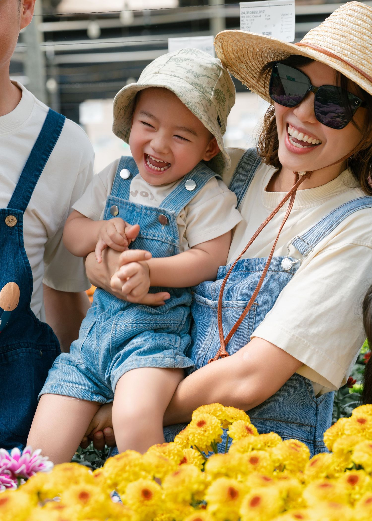 A joyful toddler in denim overalls laughs with a smiling woman wearing sunsunglasses and straw hat near yellow flowers.
