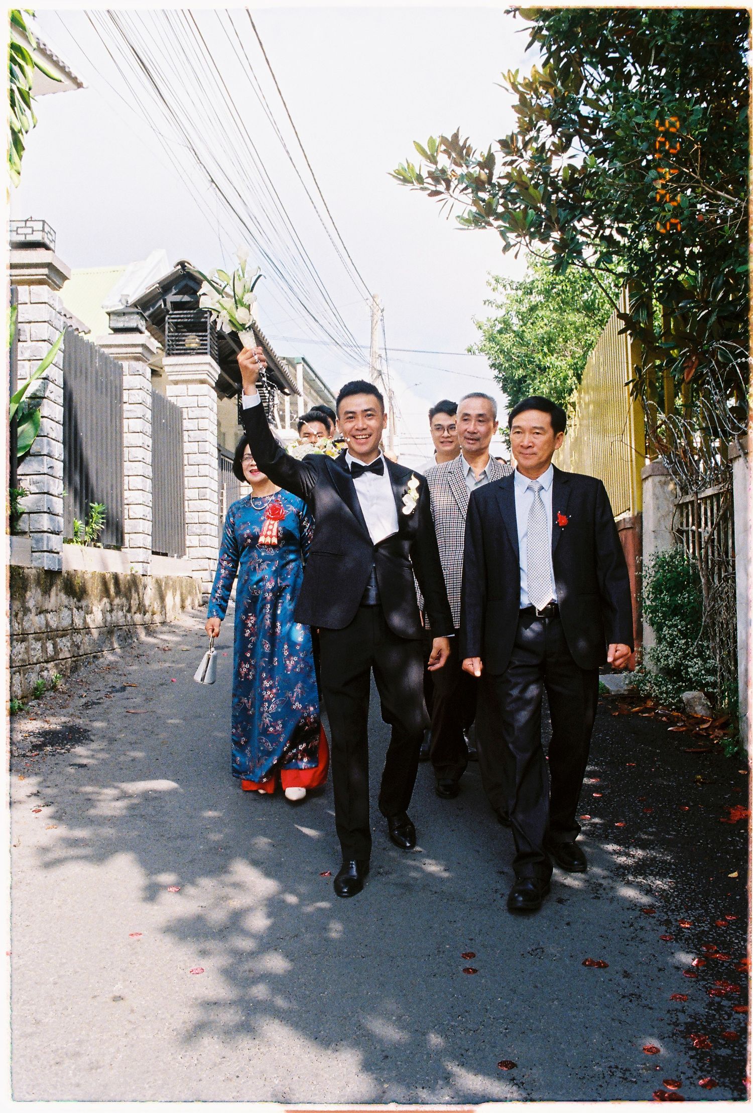 Groom in tuxedo raises flowers while walking with wedding party down a narrow street in Vietnam.