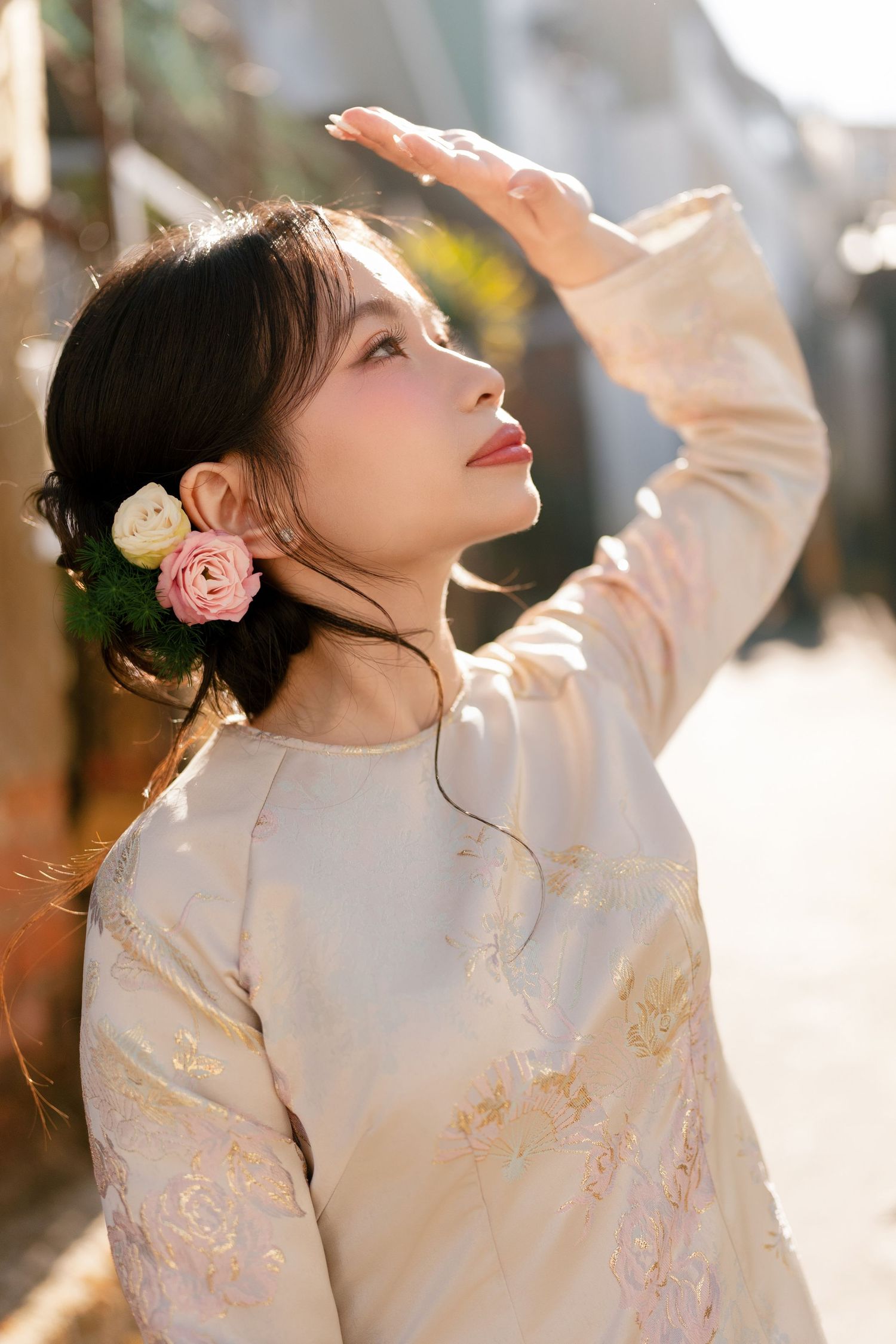 Young Asian woman in floral embroidered dress with rose hair accessory, shielding eyes from sunlight outdoors.