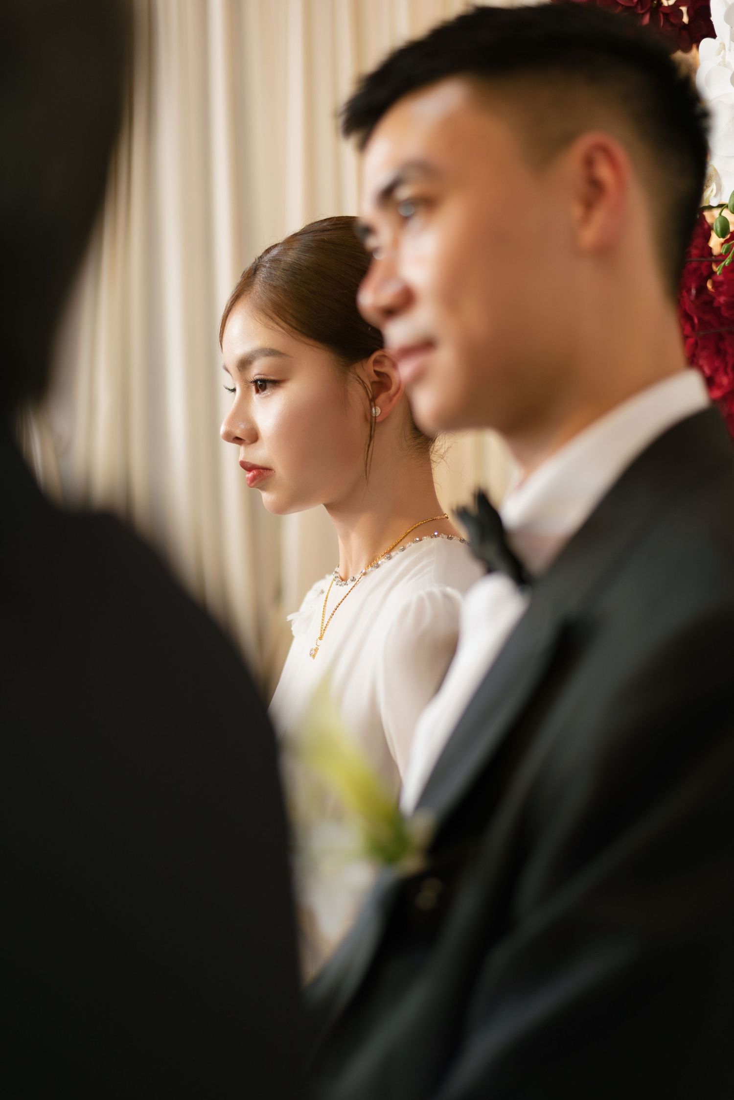 A bride in white dress and gold jewelry stands beside a groom in a black suit during a wedding ceremony.
