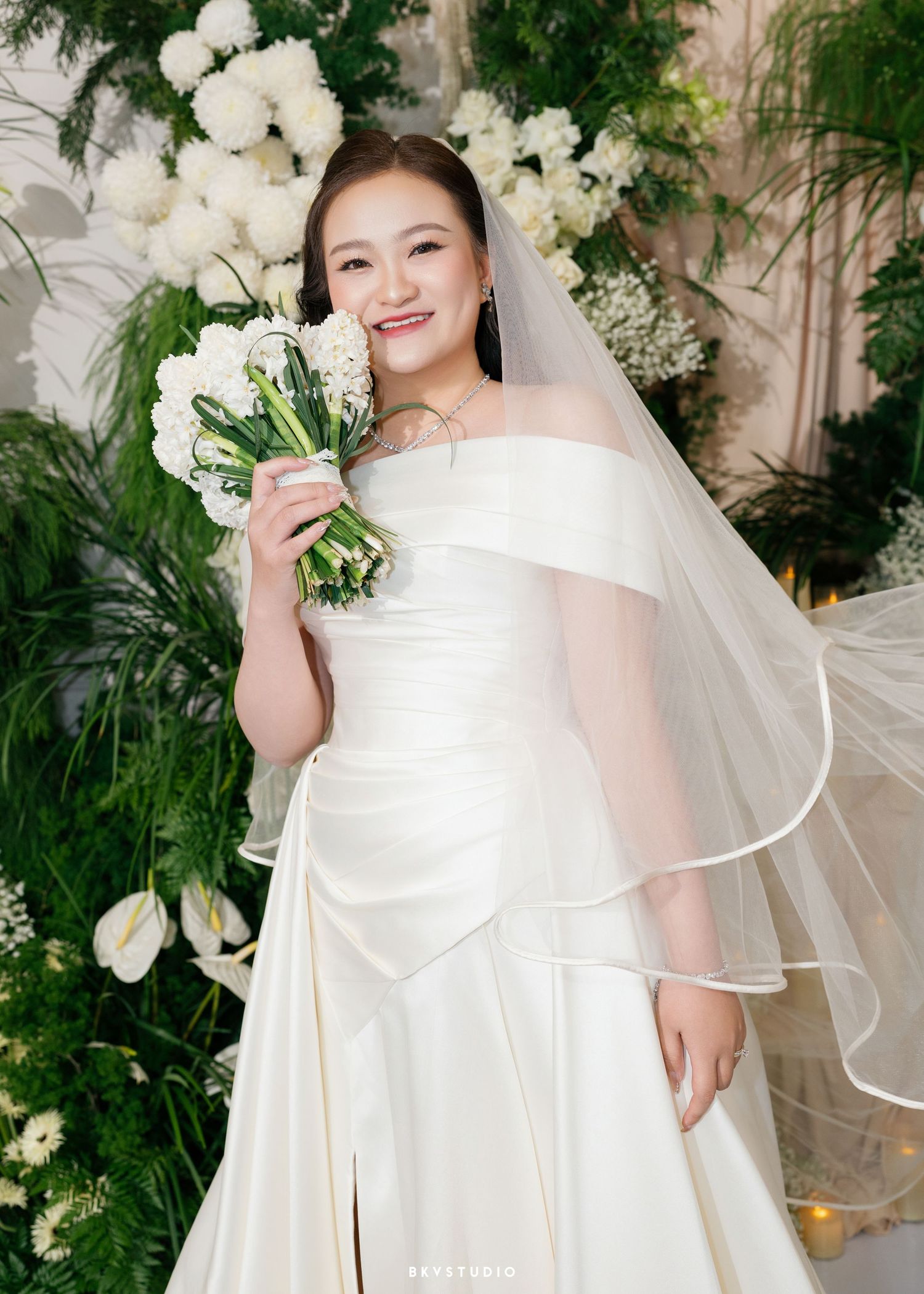 Smiling bride in white off-shoulder gown and veil holds bouquet of white flowers before floral backdrop.
