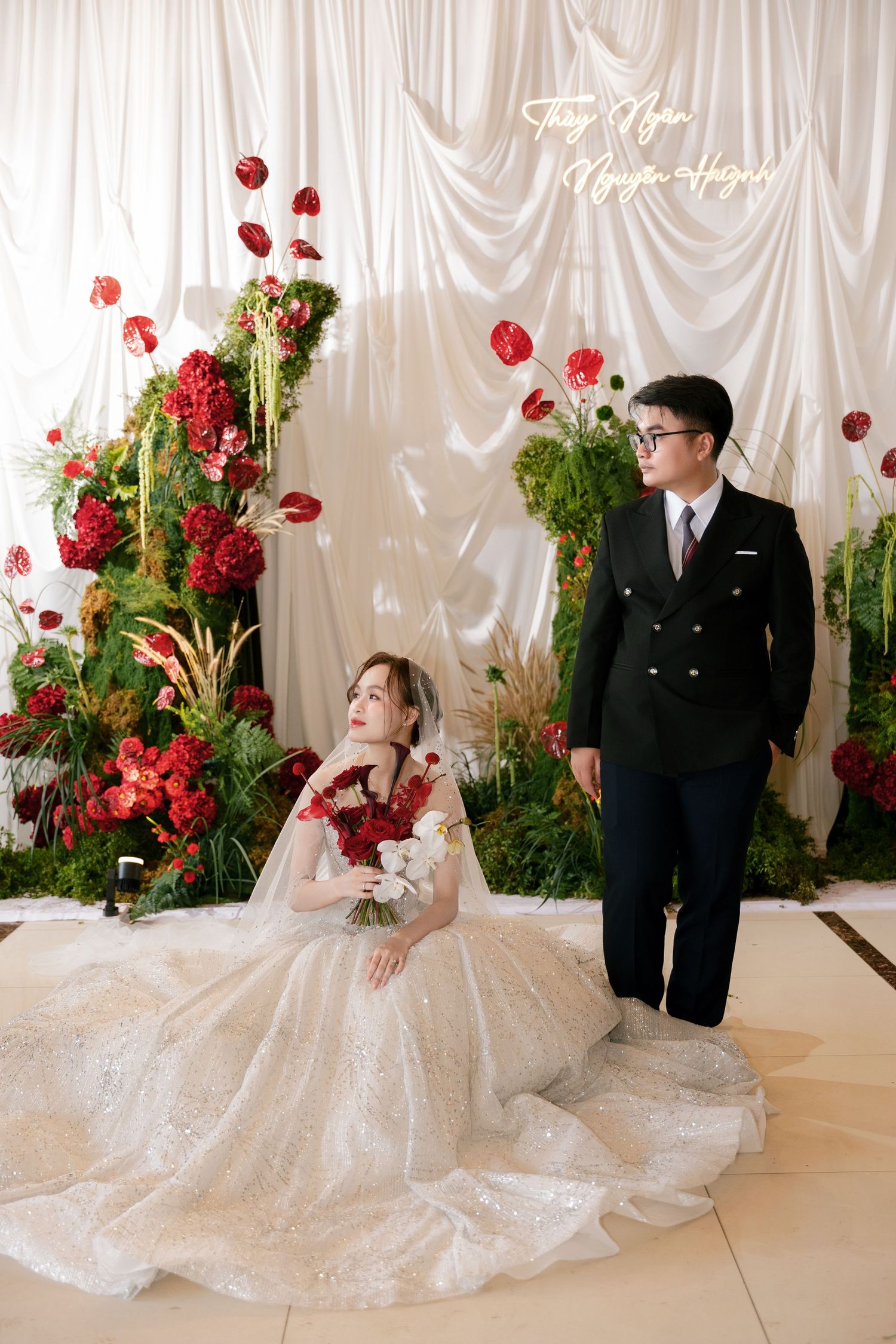 Elegant wedding photo with bride in lace gown holding red bouquet seated beside groom in black suit before floral backdrop.