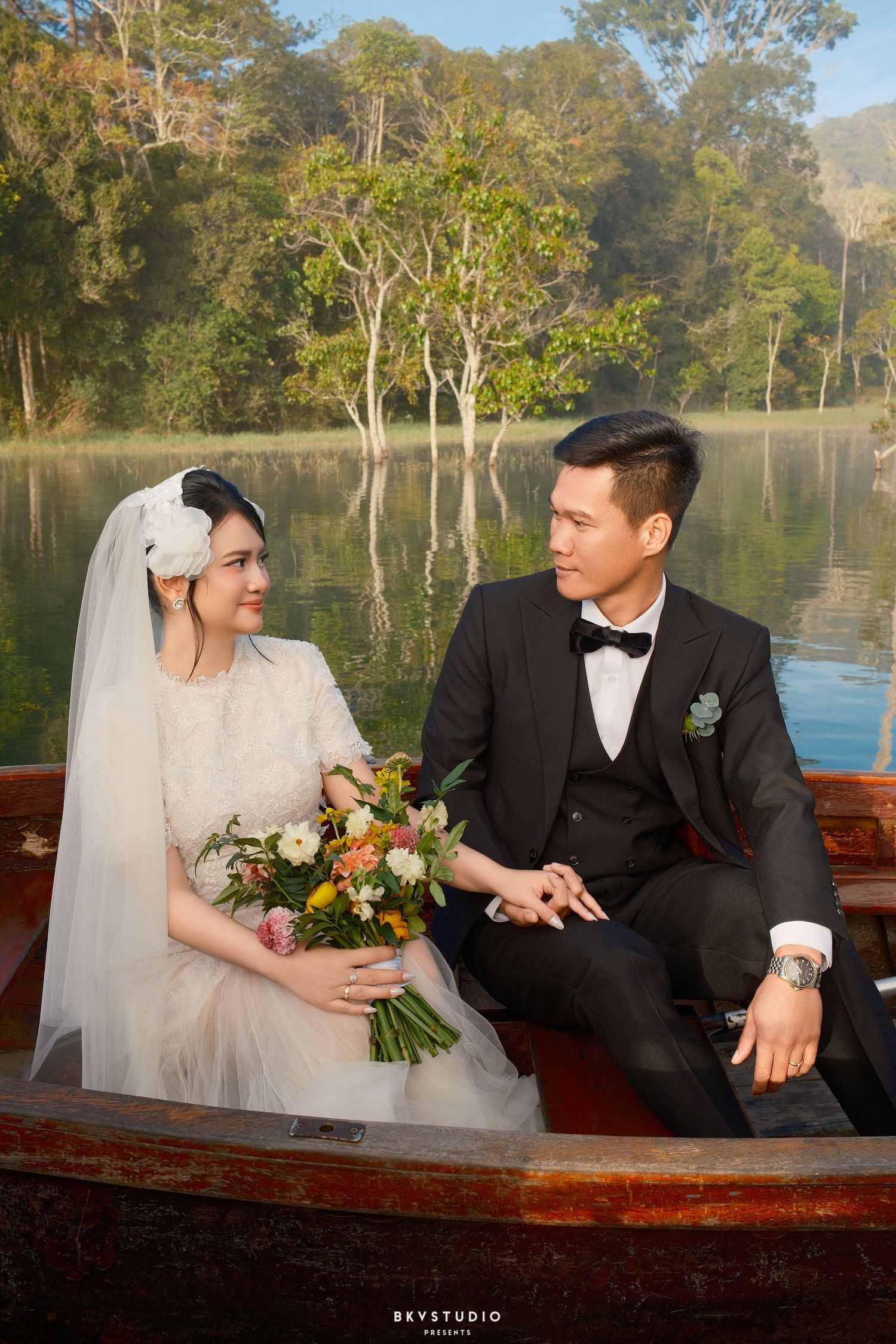 Bride and groom sitting in a wooden boat on a serene lake surrounded by lush green trees.