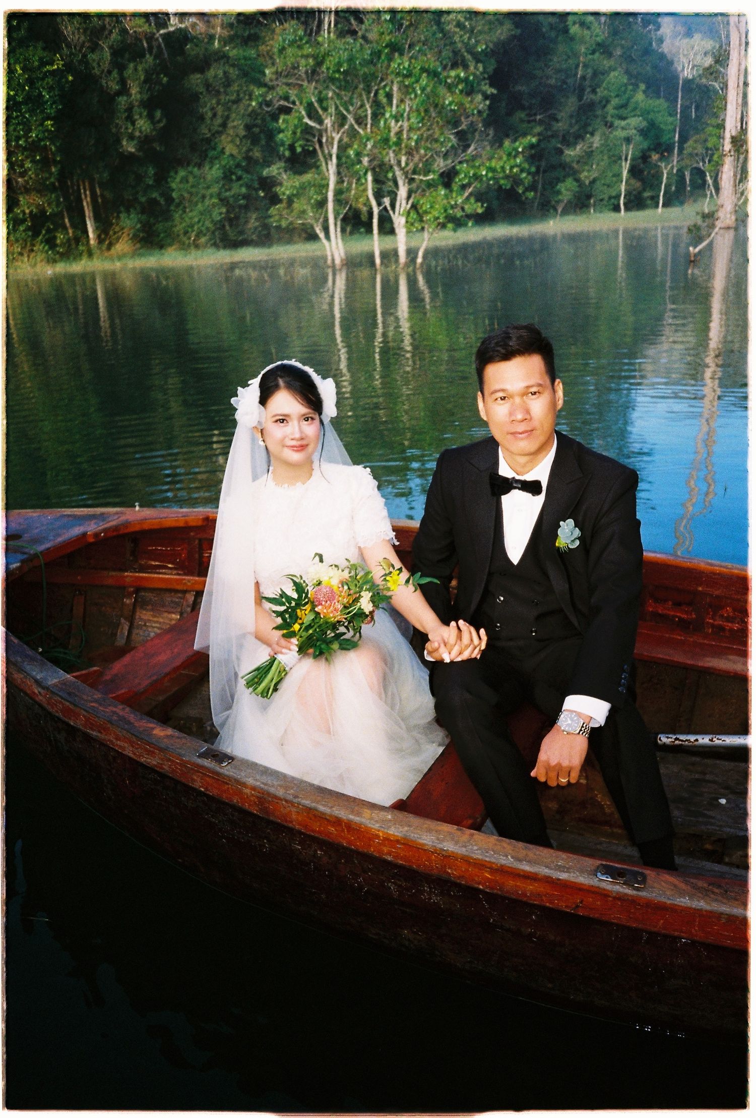 A bride and groom hold hands in a wooden boat surrounded by lush green lakeside scenery.
