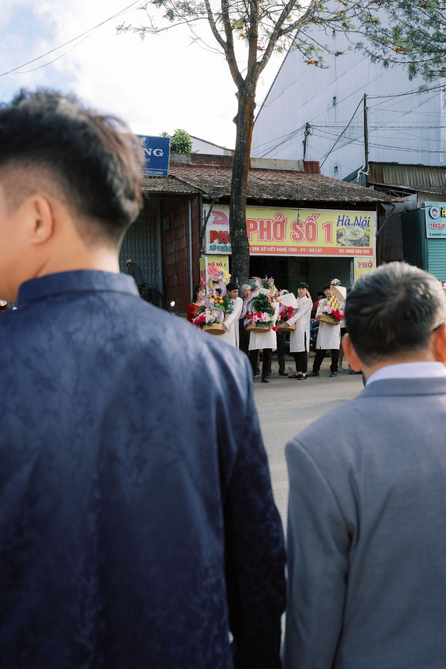 People in formal attire watching a flower ceremony outside a Vietnamese pho restaurant on a sunny day.