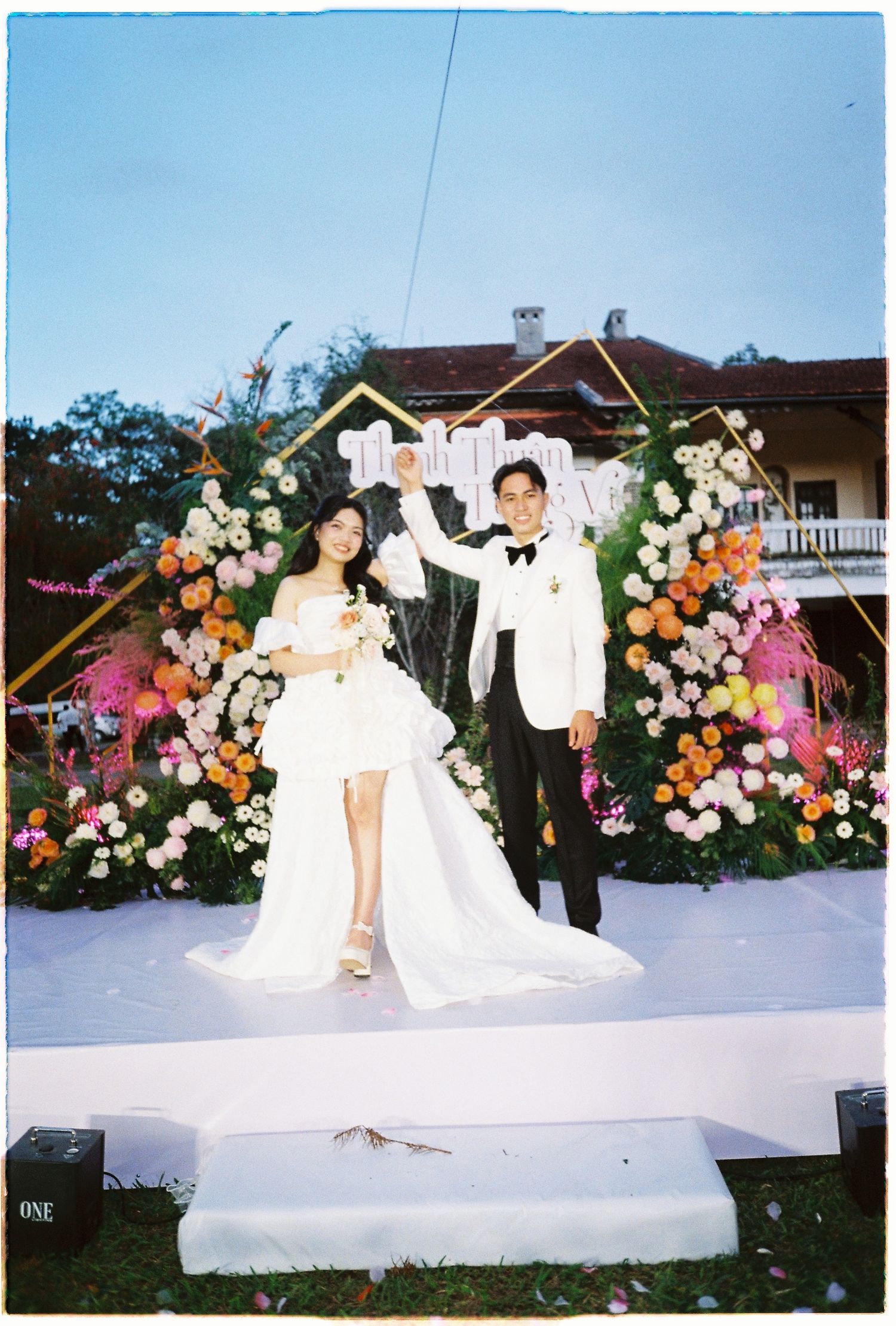 Bride and groom celebrating at outdoor wedding ceremony with colorful floral arch backdrop and white stage.