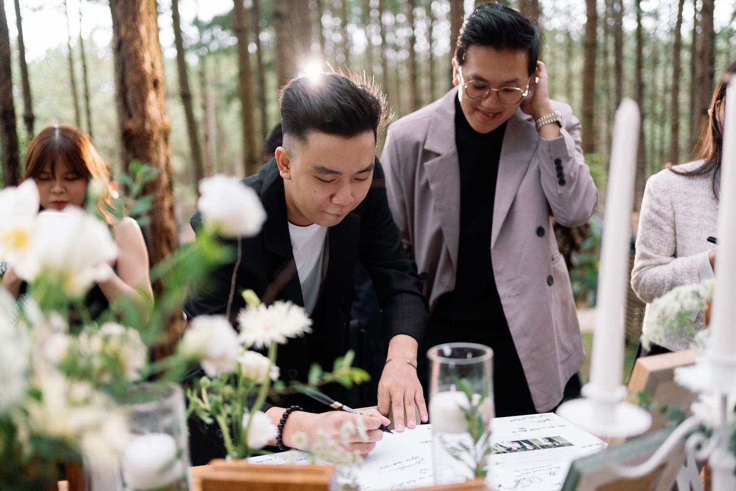 Two men in blazers signing a guest book at an outdoor wedding ceremony surrounded by white flowers.