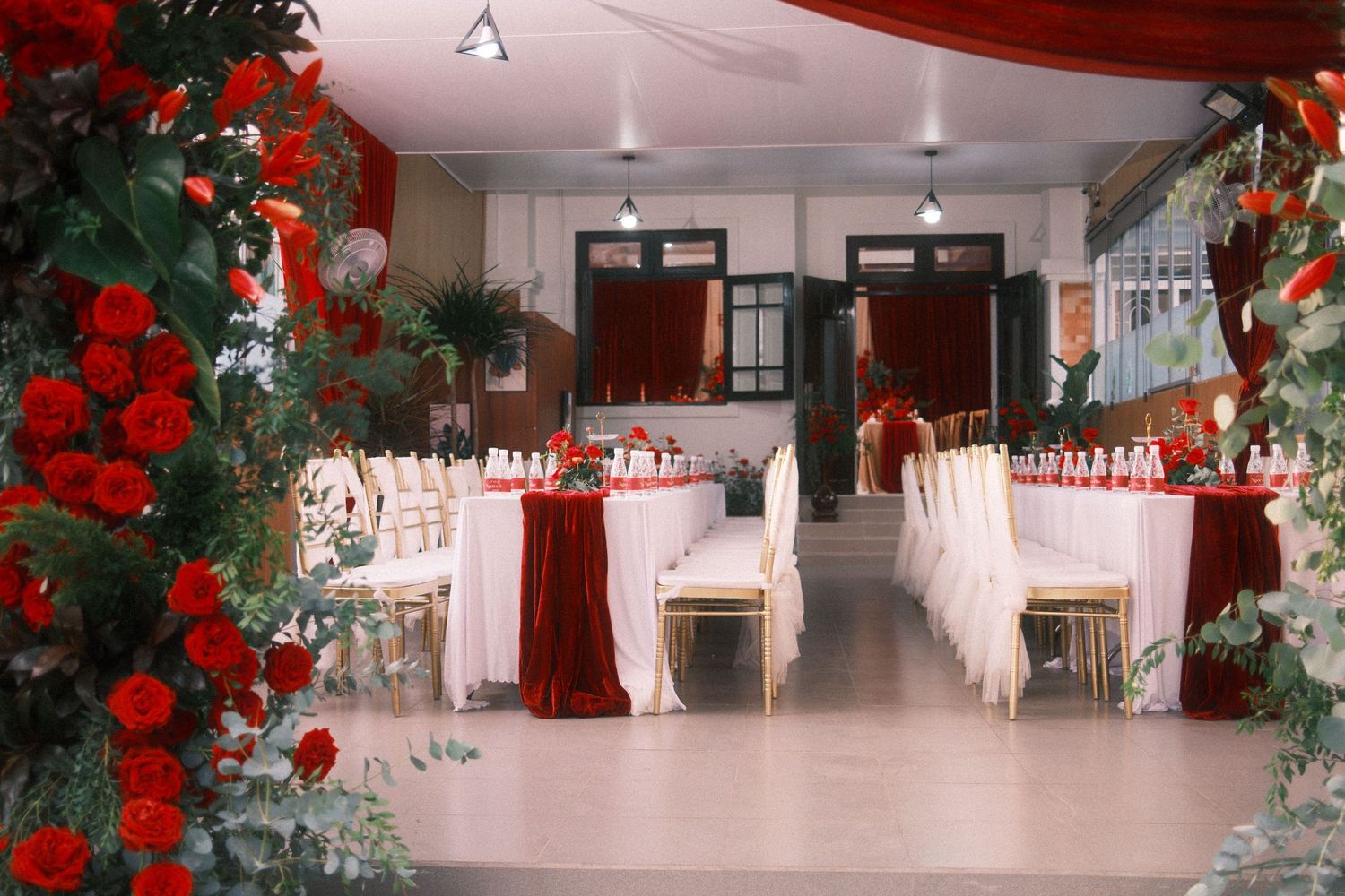 Elegant wedding reception venue decorated with red roses, white tablecloths, and gold chairs in a festive red and white theme.