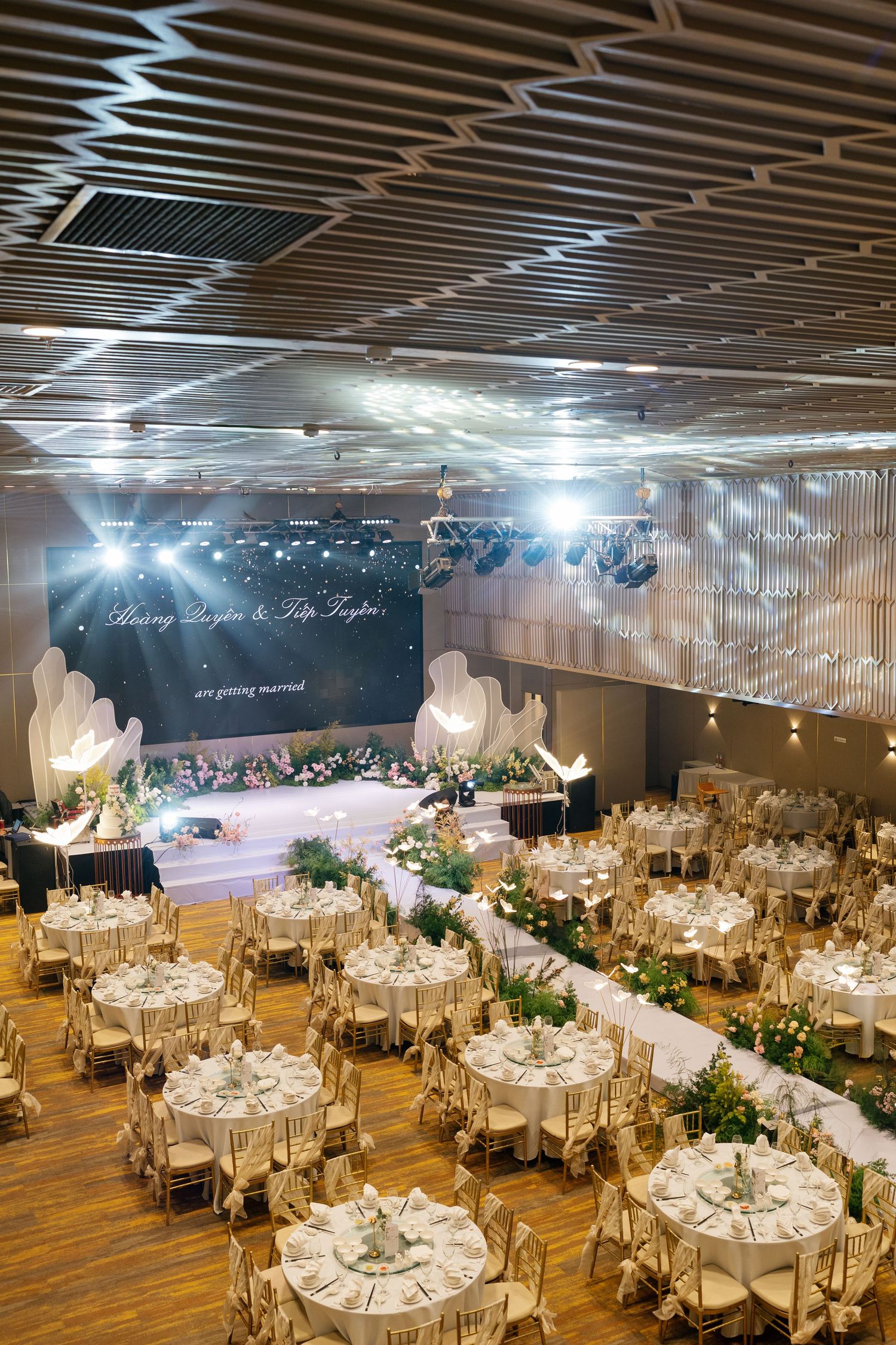 Elegant wedding reception hall with round tables, floral centerpieces, gold chairs, and illuminated stage backdrop.