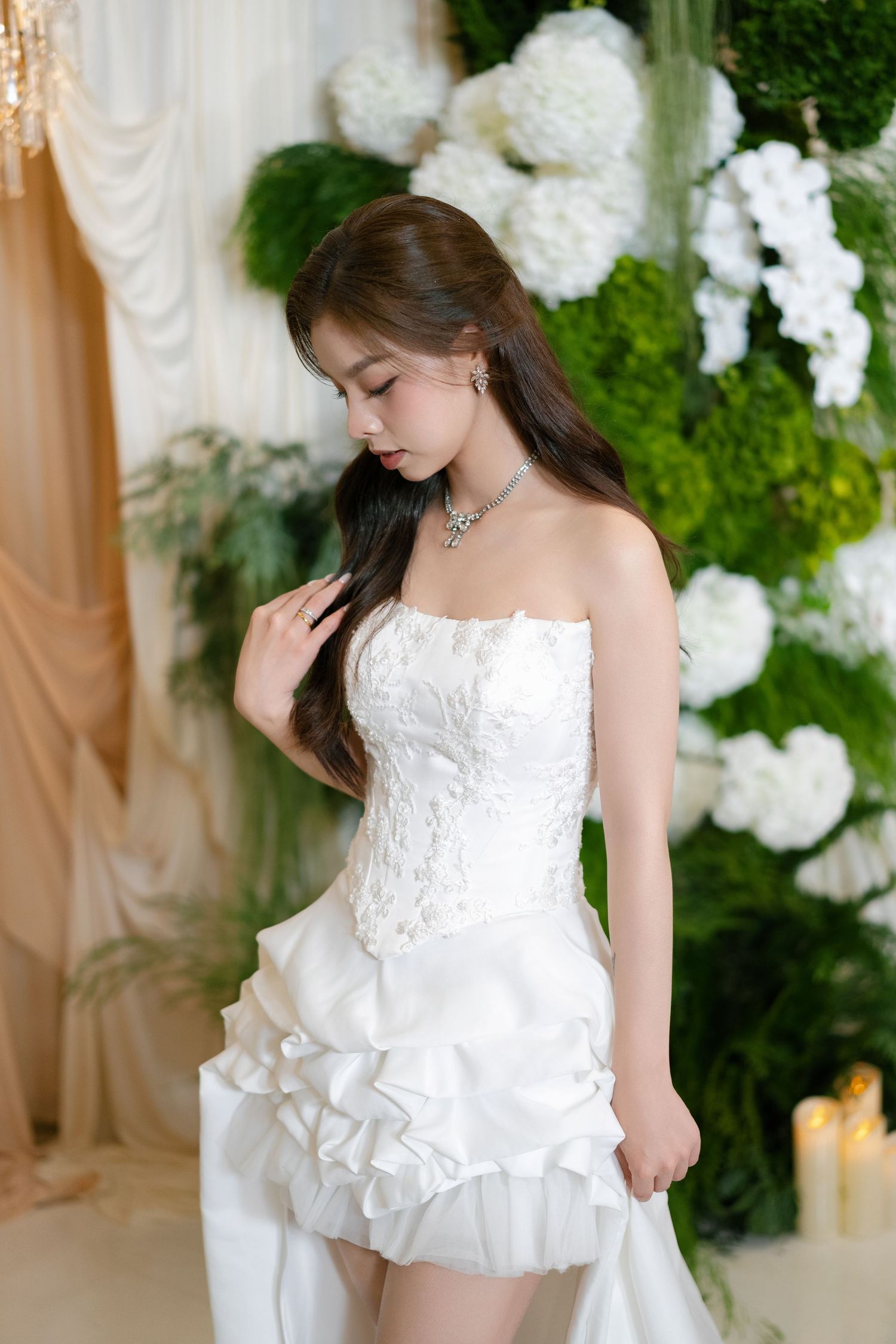 Young woman in a white strapless bridal mini dress with lace, wearing a diamond necklace, posing before floral backdrop.