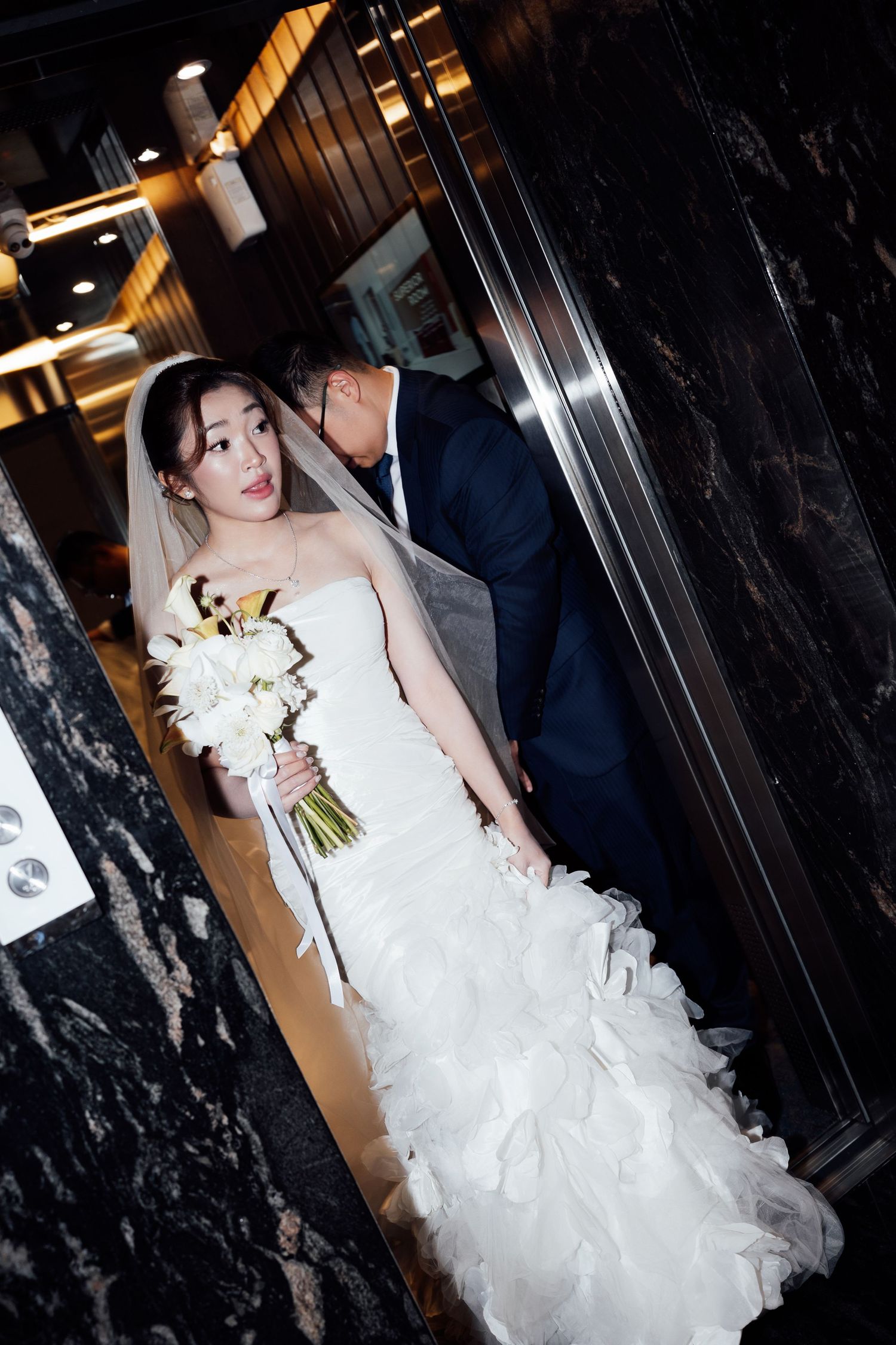 Bride in white strapless ruffled gown holding white bouquet while groom in navy suit kisses her cheek in elegant venue.
