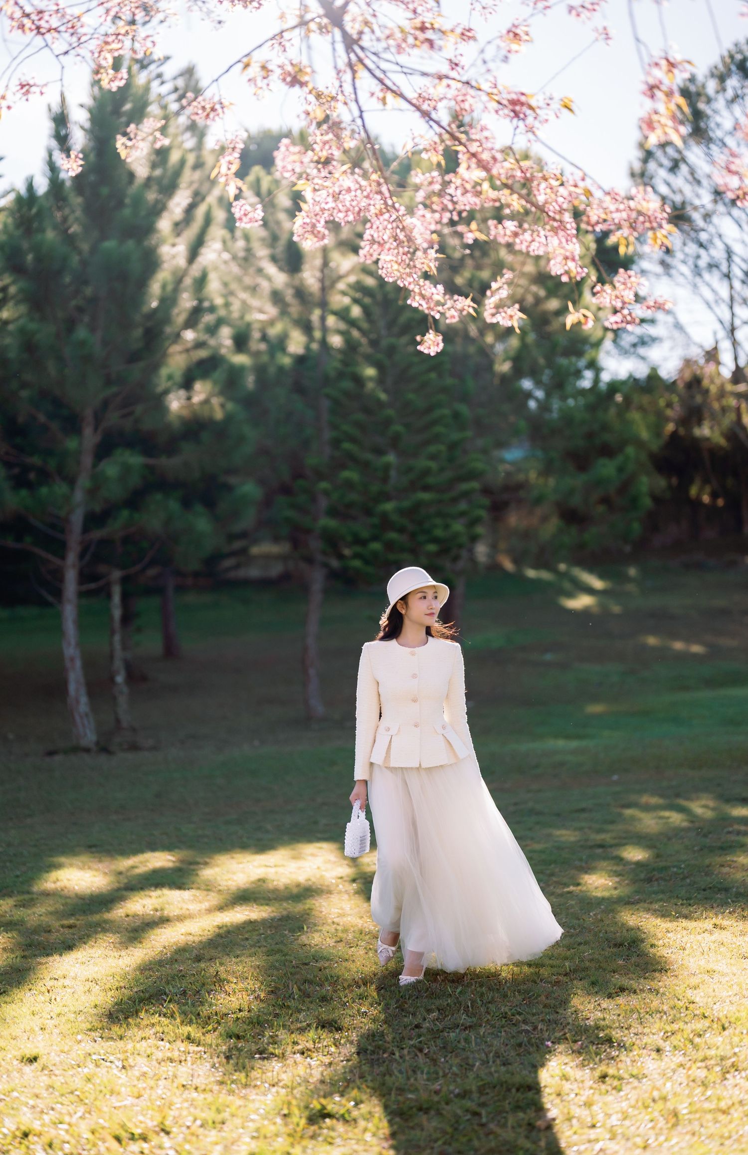 Woman in cream outfit and hat stands under pink cherry blossoms in a sunlit green park.