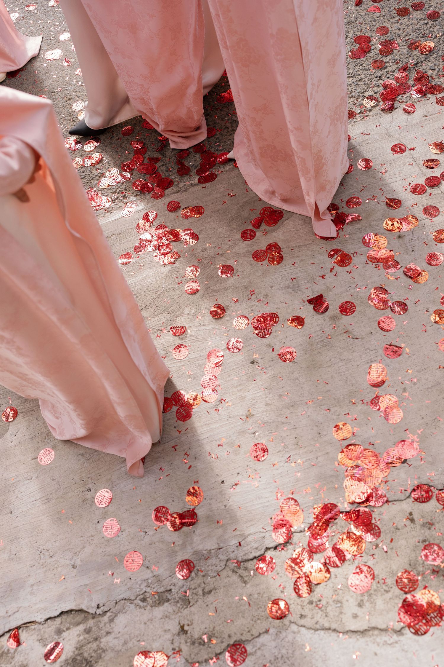 Bridesmaids in blush pink gowns stand on concrete floor scattered with red and pink rose petals at a wedding.