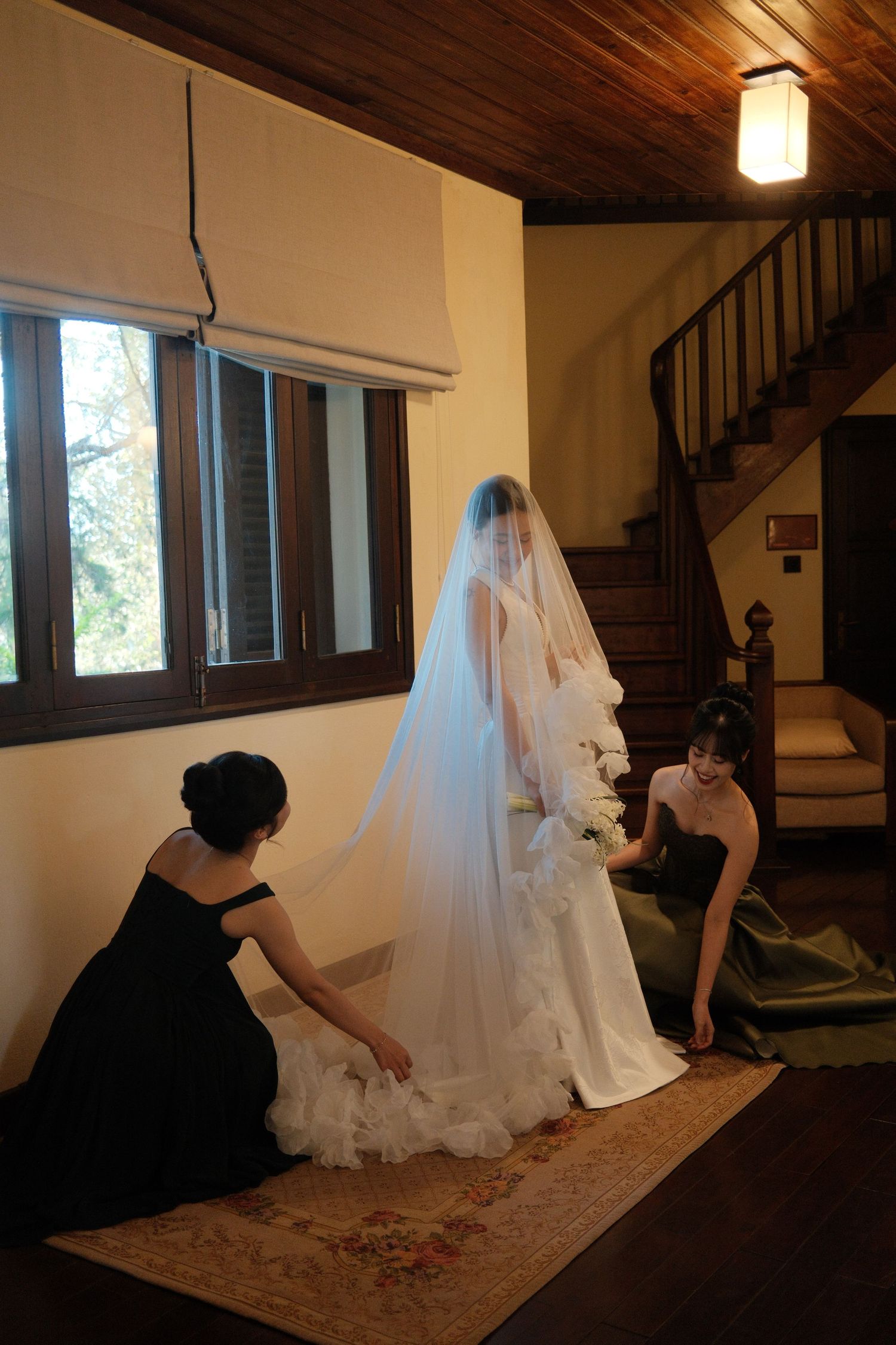 Two bridesmaids in black dresses adjust the train of a veiled bride near a wooden staircase.