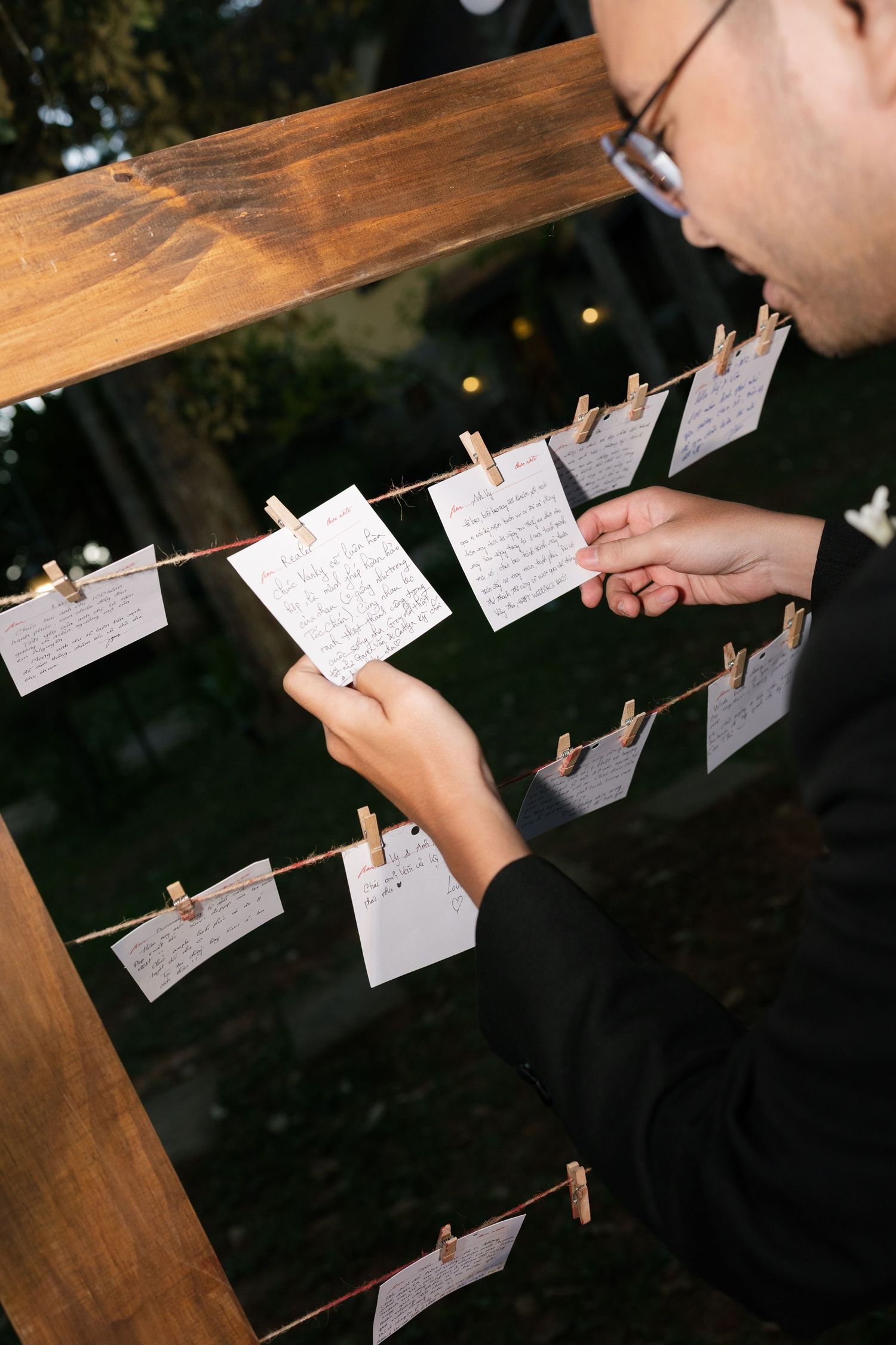 Wedding guest reading handwritten wish cards clipped to a wooden display frame at outdoor reception.