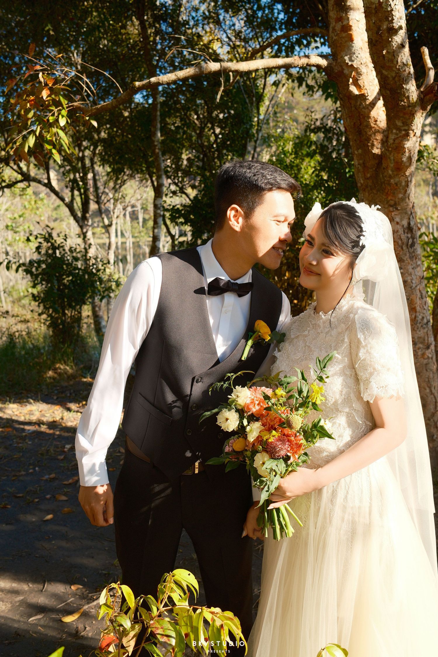 Newlyweds share a tender kiss outdoors, bride holding orange and white floral bouquet, groom in black vest and bow tie.