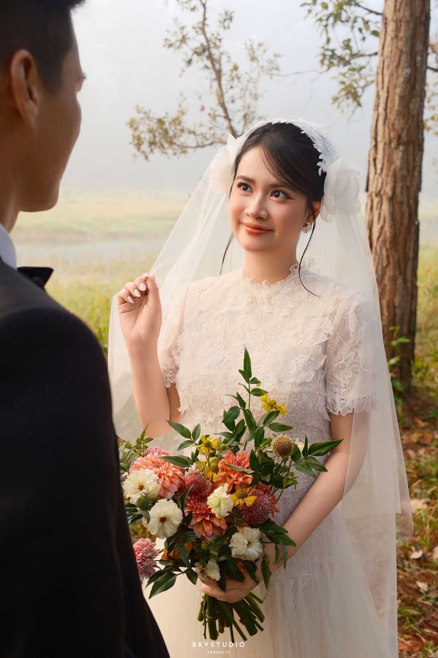 Smiling bride in lace wedding dress and veil holds colorful bouquet outdoors in misty natural setting.