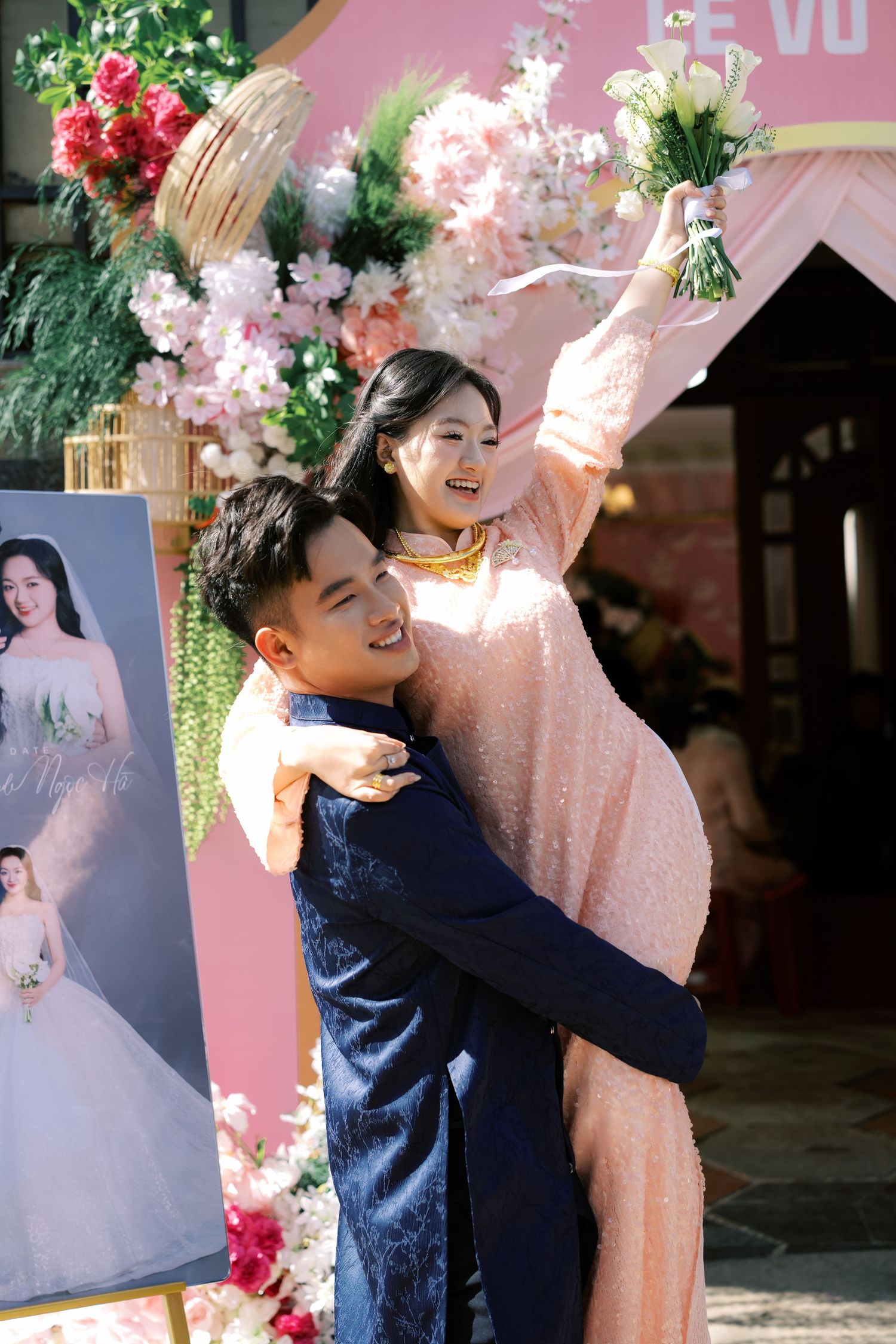 Joyful Vietnamese bride in pink sequined ao dai is lifted by groom in blue ao dai at their wedding.
