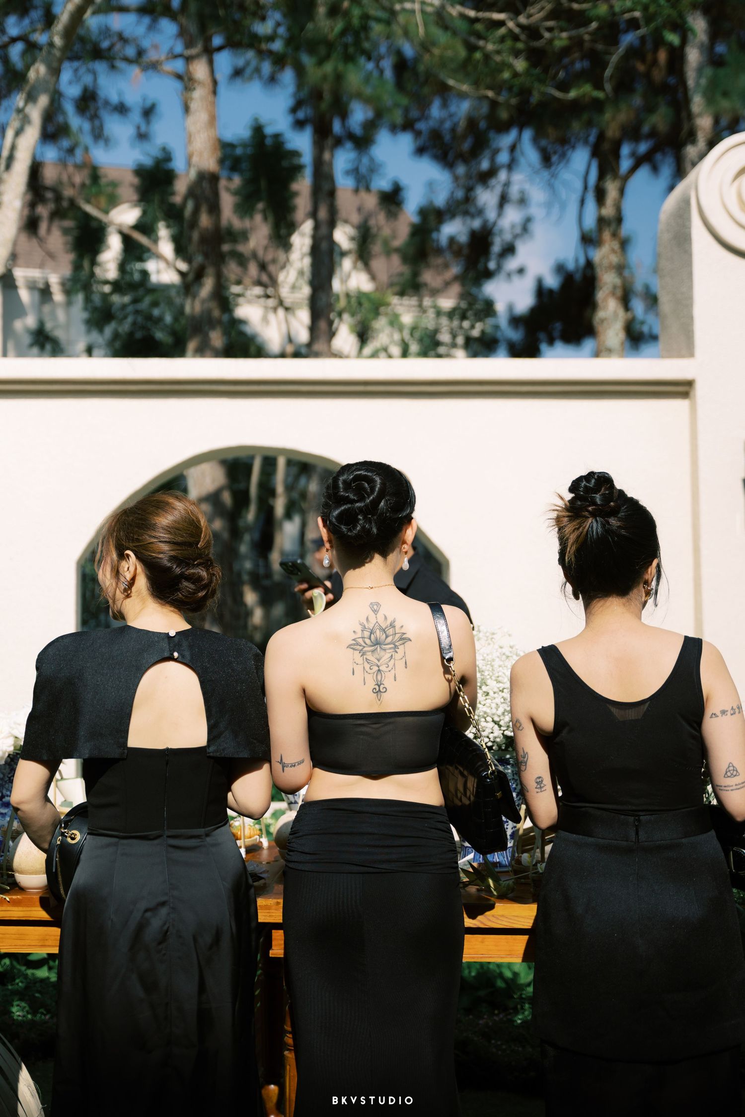Three bridesmaids in black dresses viewed from behind, one with ornate back tattoo at wedding.
