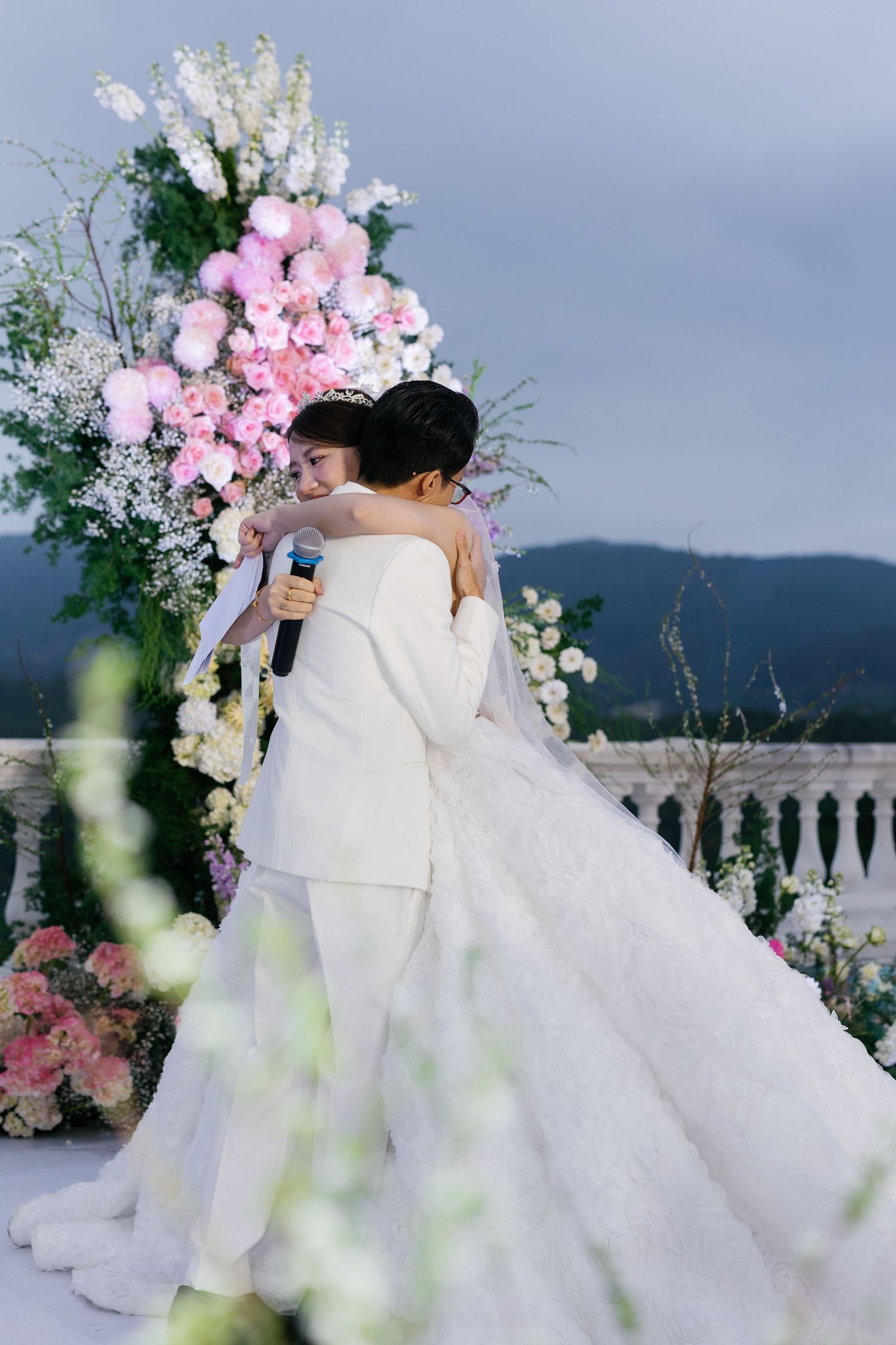 Bride and groom embracing at outdoor wedding ceremony with pink floral arch and mountain backdrop.