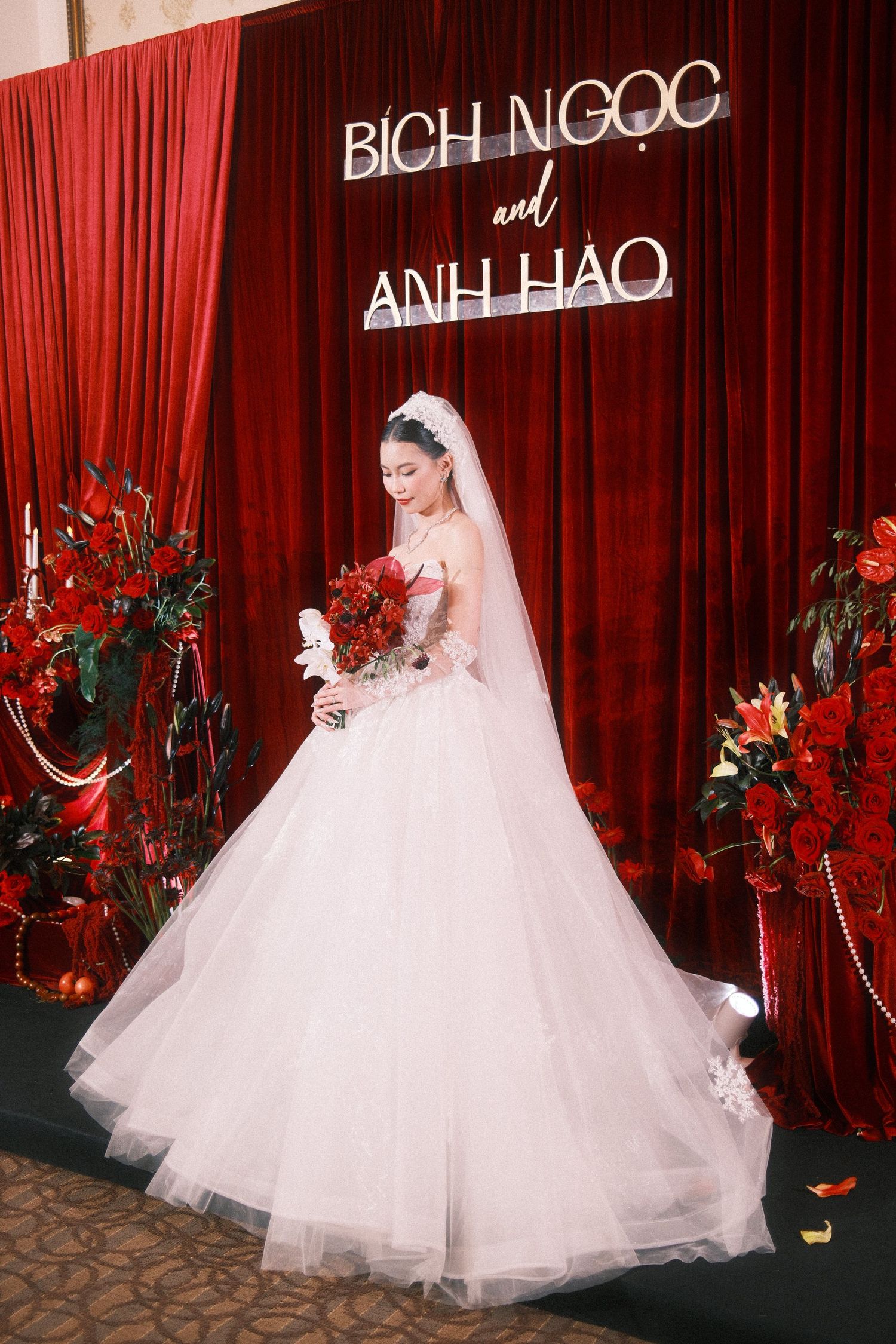 Bride in white ball gown holds red bouquet before a red floral backdrop at Bich Ngoc and Anh Hao wedding.