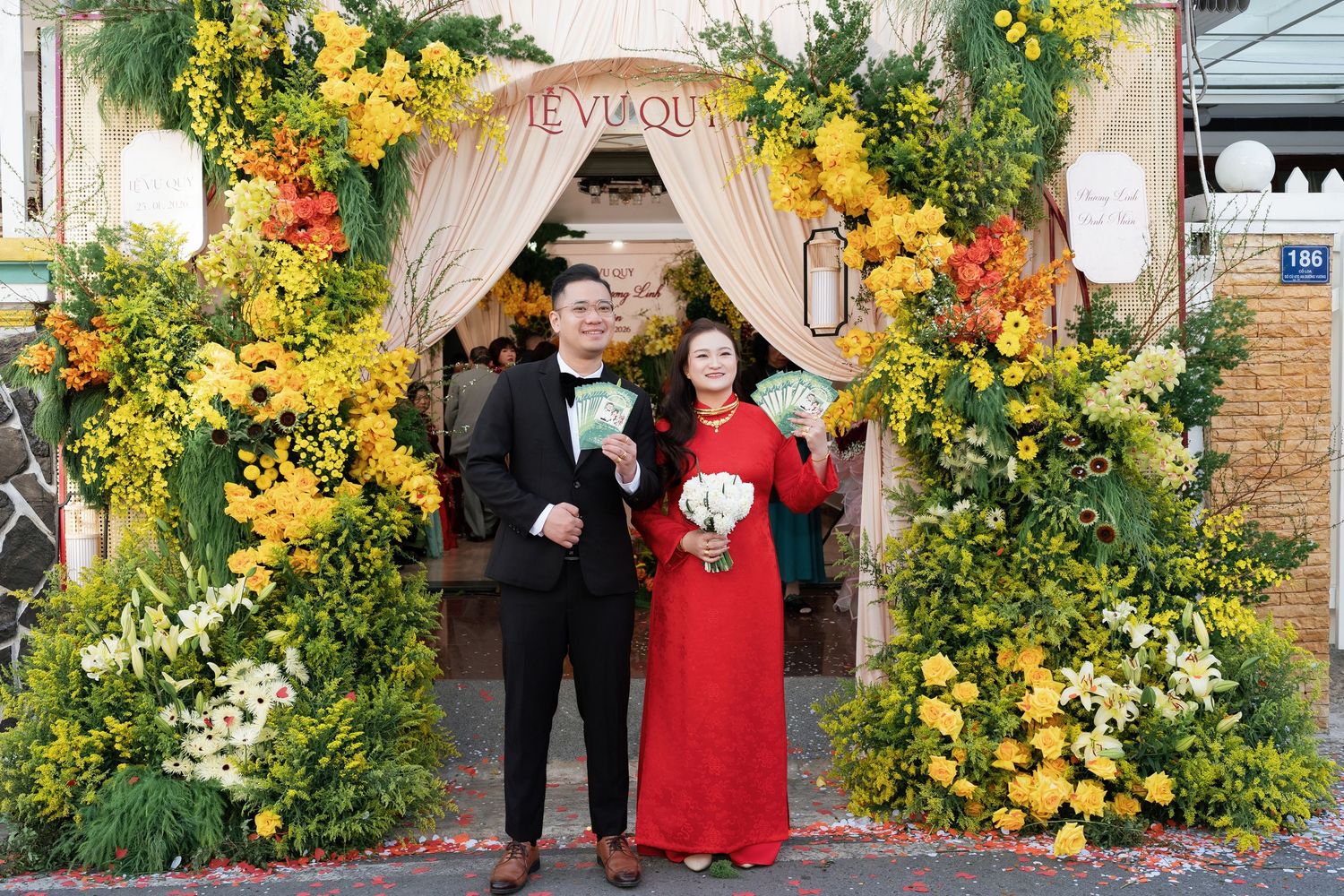 Couple in traditional Vietnamese wedding attire posing before a vibrant yellow floral arch entrance decoration.