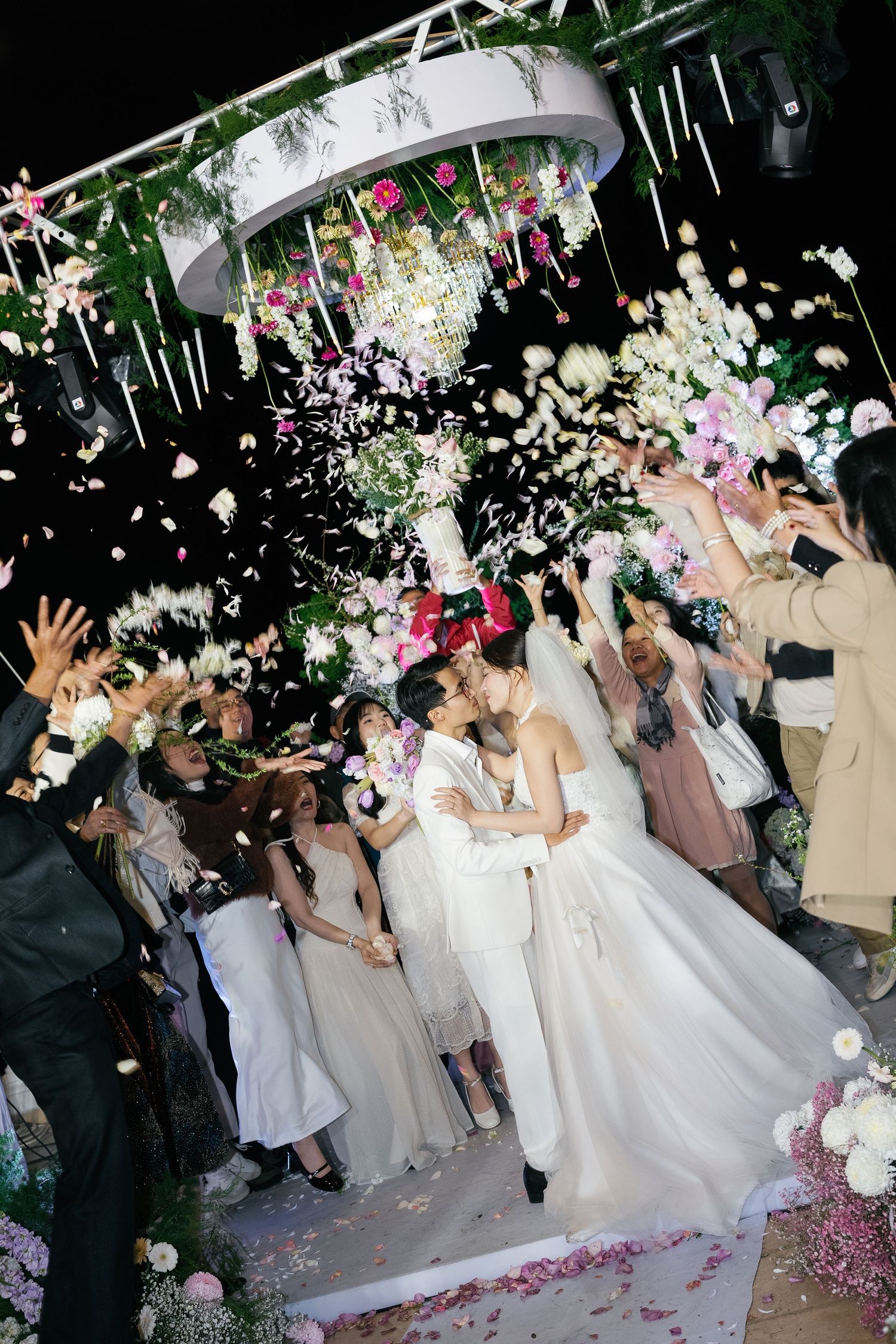 Bride and groom share a kiss at outdoor wedding ceremony as guests toss flower petals under chandelier.