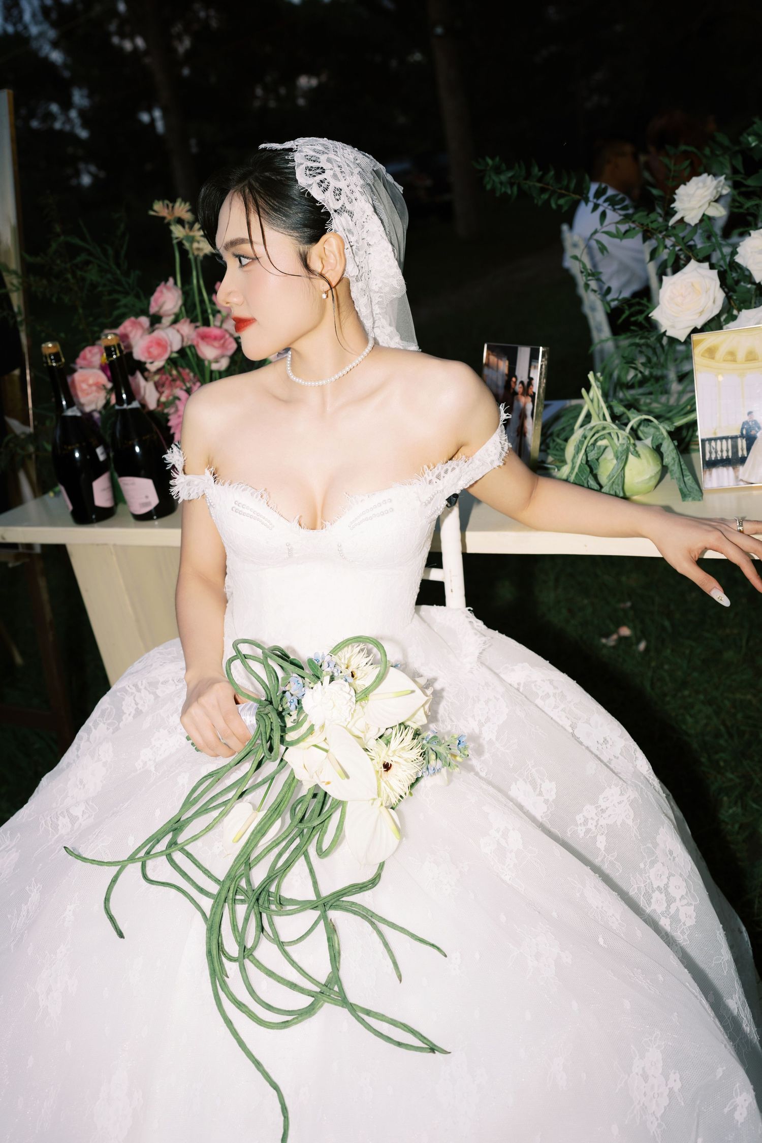 Bride in off-shoulder white ball gown with lace veil holds bouquet of white flowers with trailing green stems outdoors.