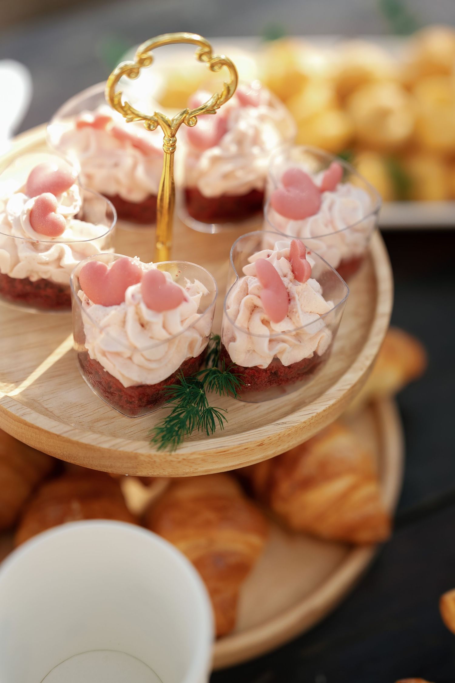 Elegant two-tier wooden stand with pink cream dessert cups topped with heart decorations, surrounded by croissants.
