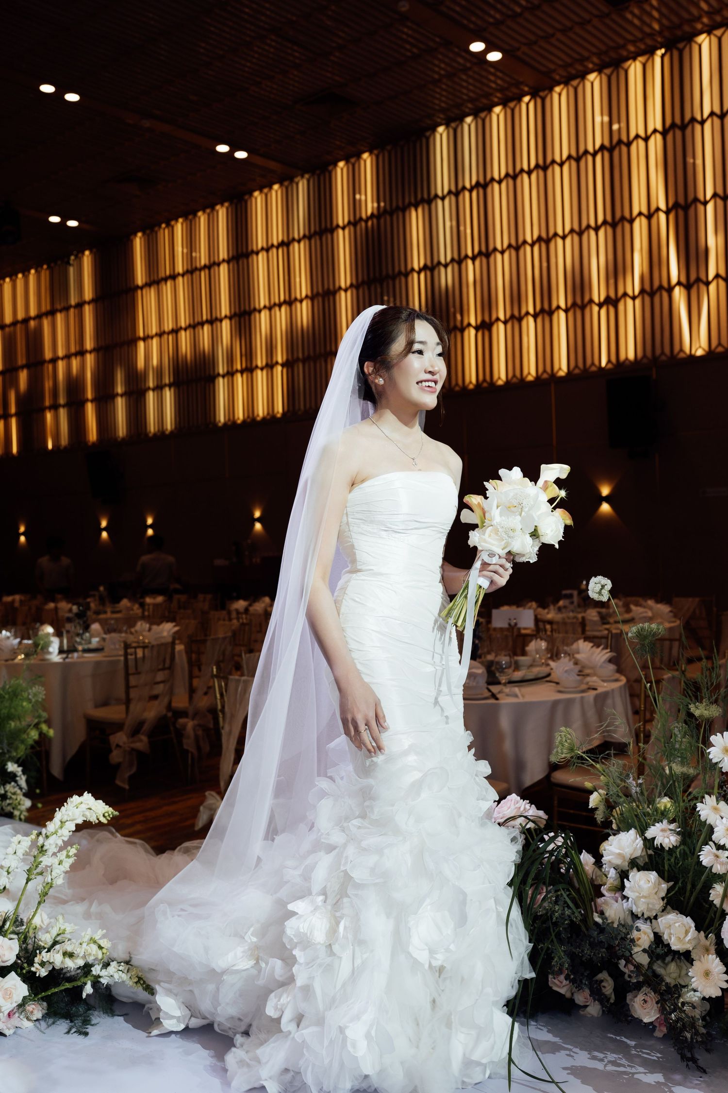 Smiling Asian bride in strapless white ruffle gown and veil holds white bouquet in elegant wedding reception venue.