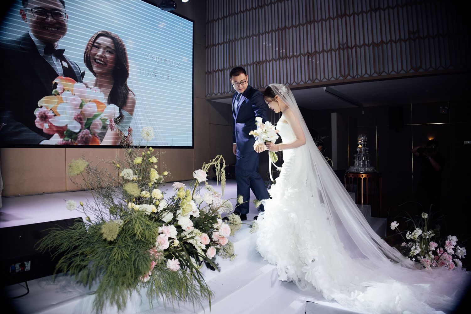 Bride in white gown and groom stand at elegant wedding ceremony with floral arrangements and projection screen.