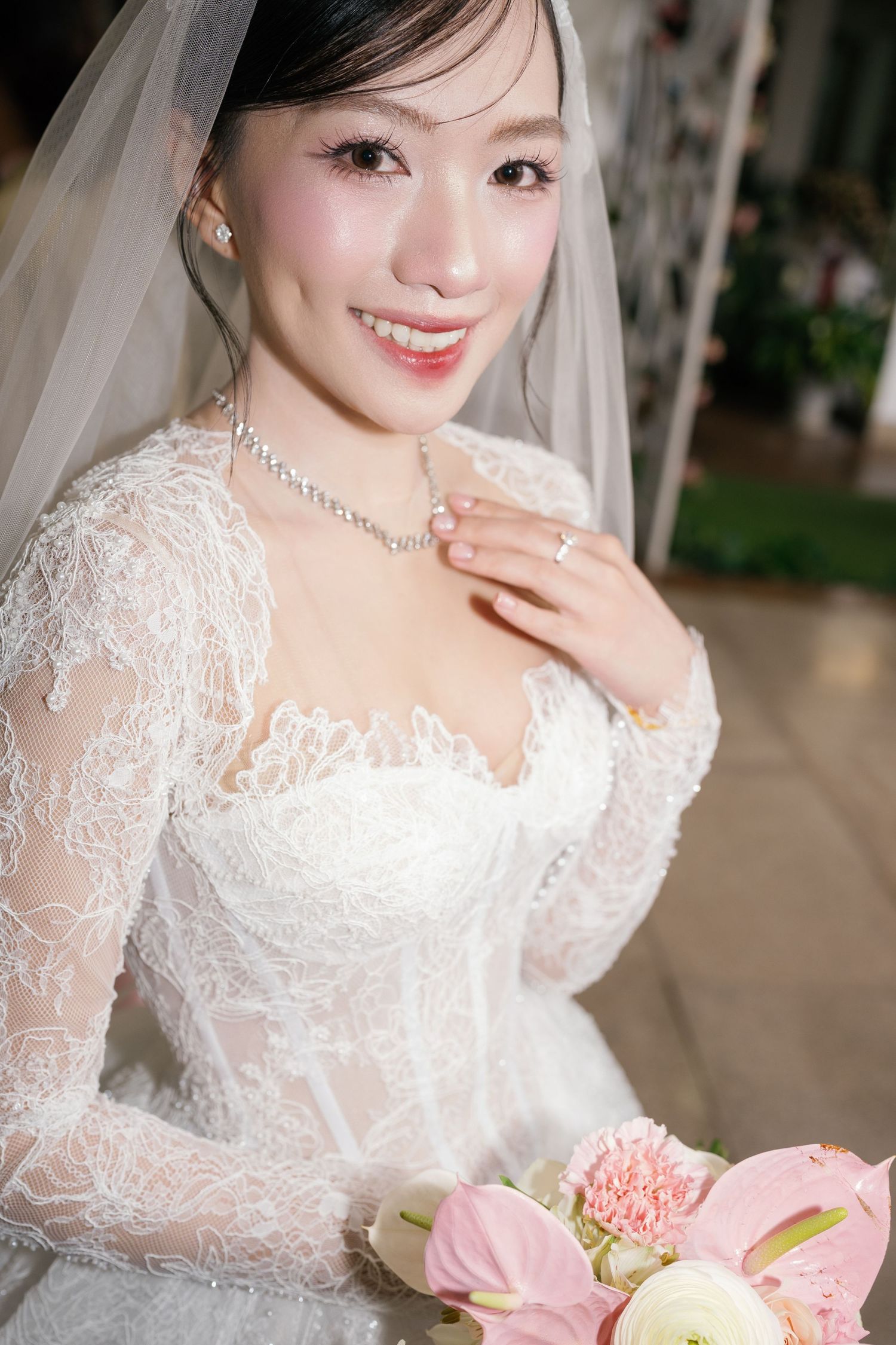 Smiling Asian bride in lace wedding dress with veil holds pink bouquet and diamond necklace at indoor ceremony.