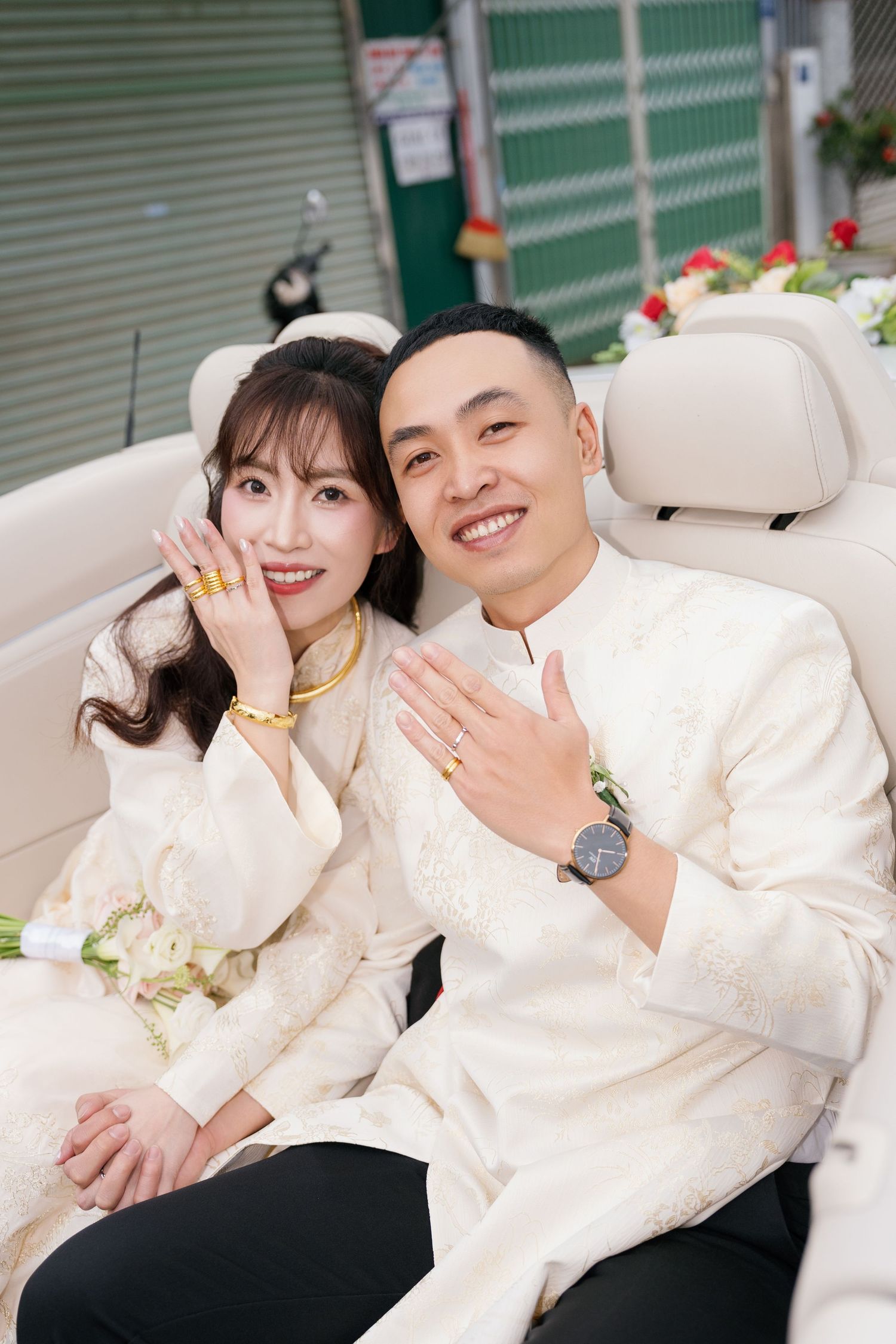 Vietnamese couple in traditional white ao dai wedding attire sitting in car, showing gold jewelry and rings.