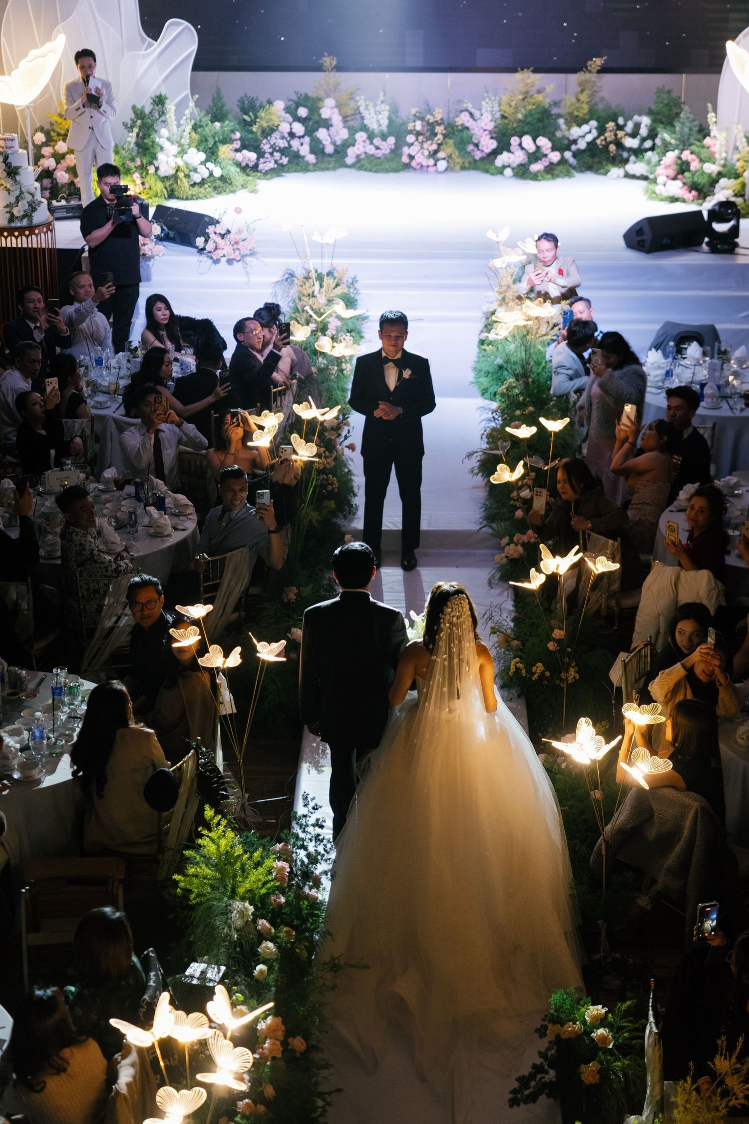 Aerial view of bride and groom walking down a stunning wedding aisle with glowing butterfly lights and lush greenery.
