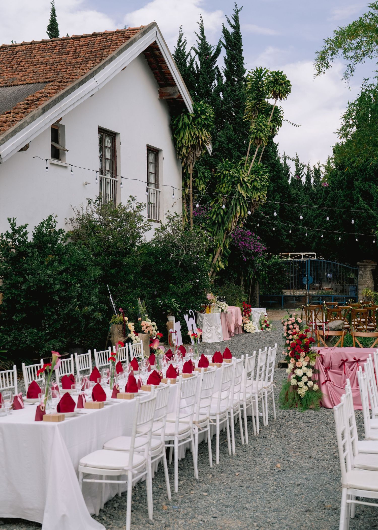 Elegant outdoor wedding reception with long white tables, red napkins, floral centerpieces, and string lights near a white building.