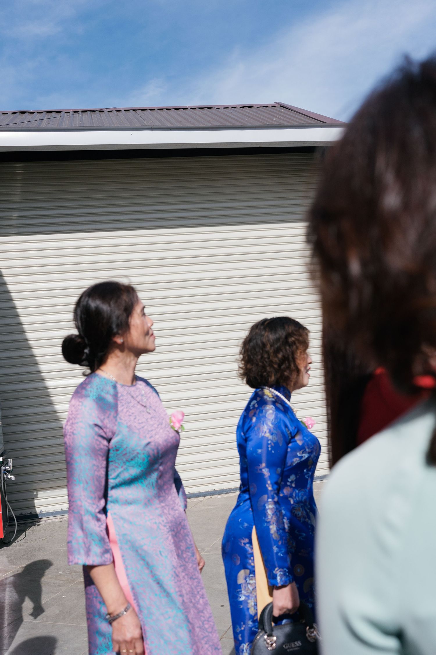 Two women in colorful ao dai dresses stand outdoors in bright sunlight near a metal building.