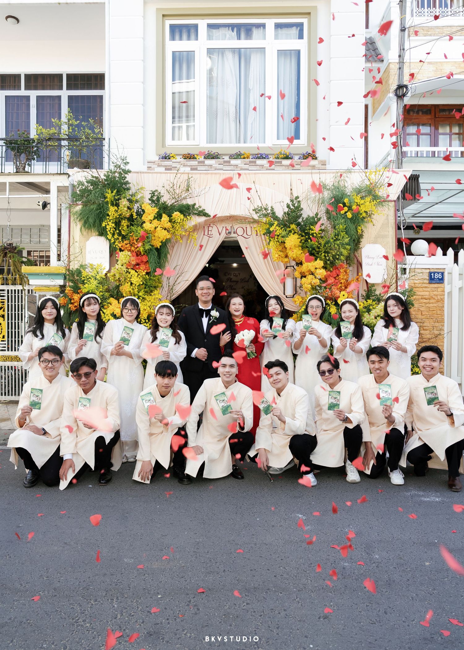 Large Vietnamese wedding party in white ao dai attire poses outdoors with red flower petals falling.