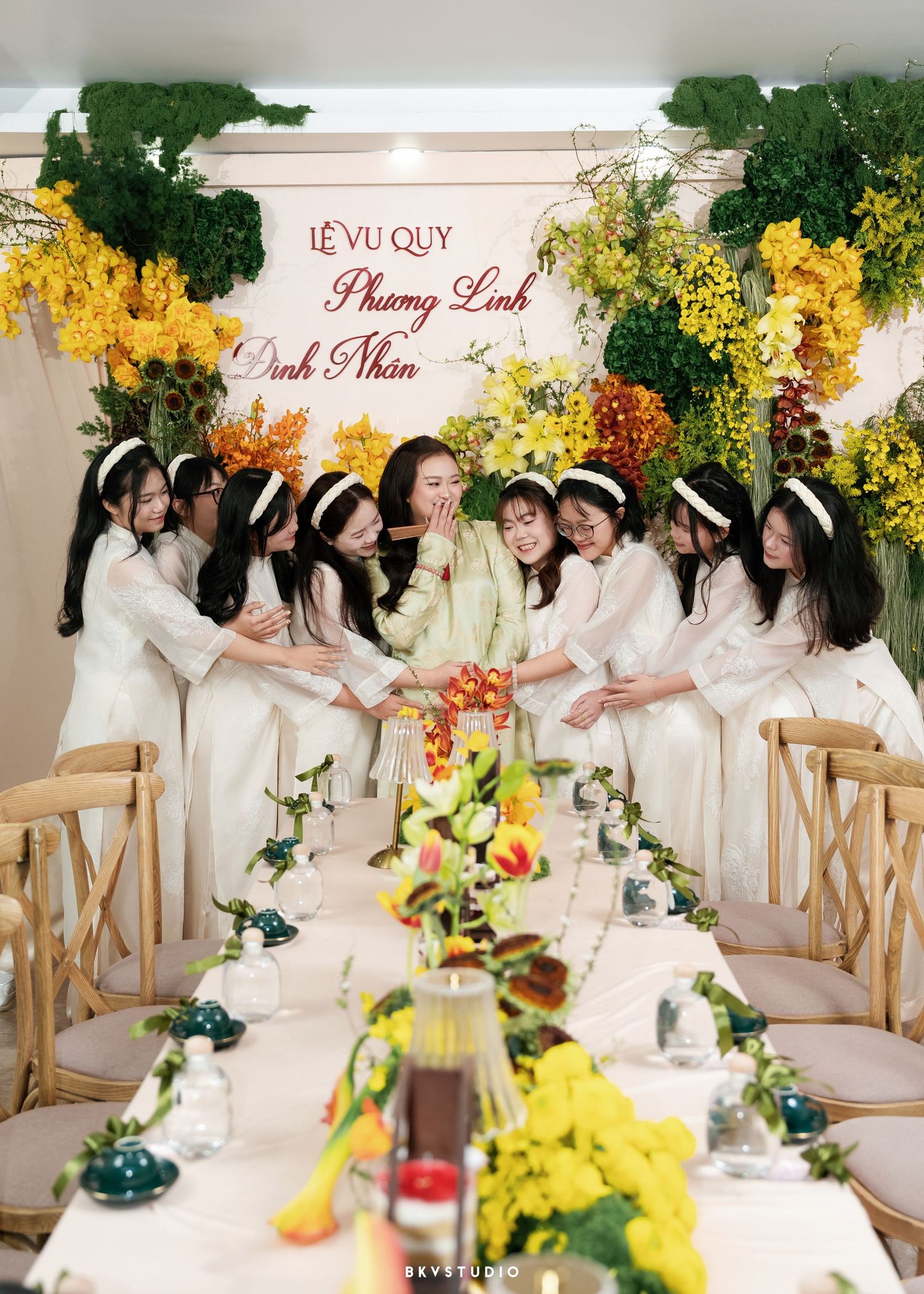 Vietnamese wedding ceremony with bridesmaids in white ao dai gathered around bride under floral arch at Le Vu Quy celebration.