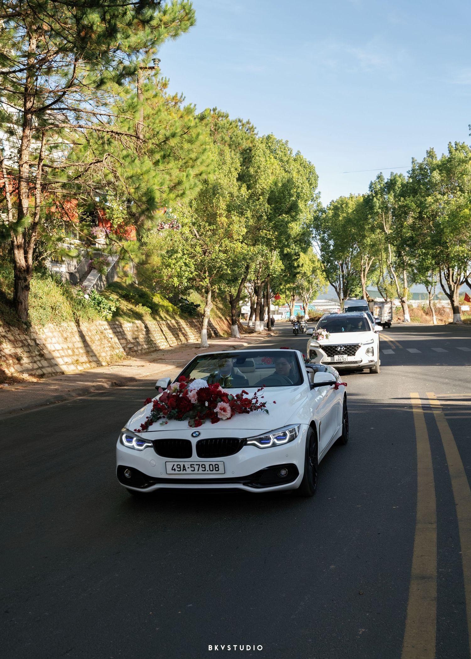 White BMW convertible decorated with red flowers leads a wedding convoy down a tree-lined road in Vietnam.