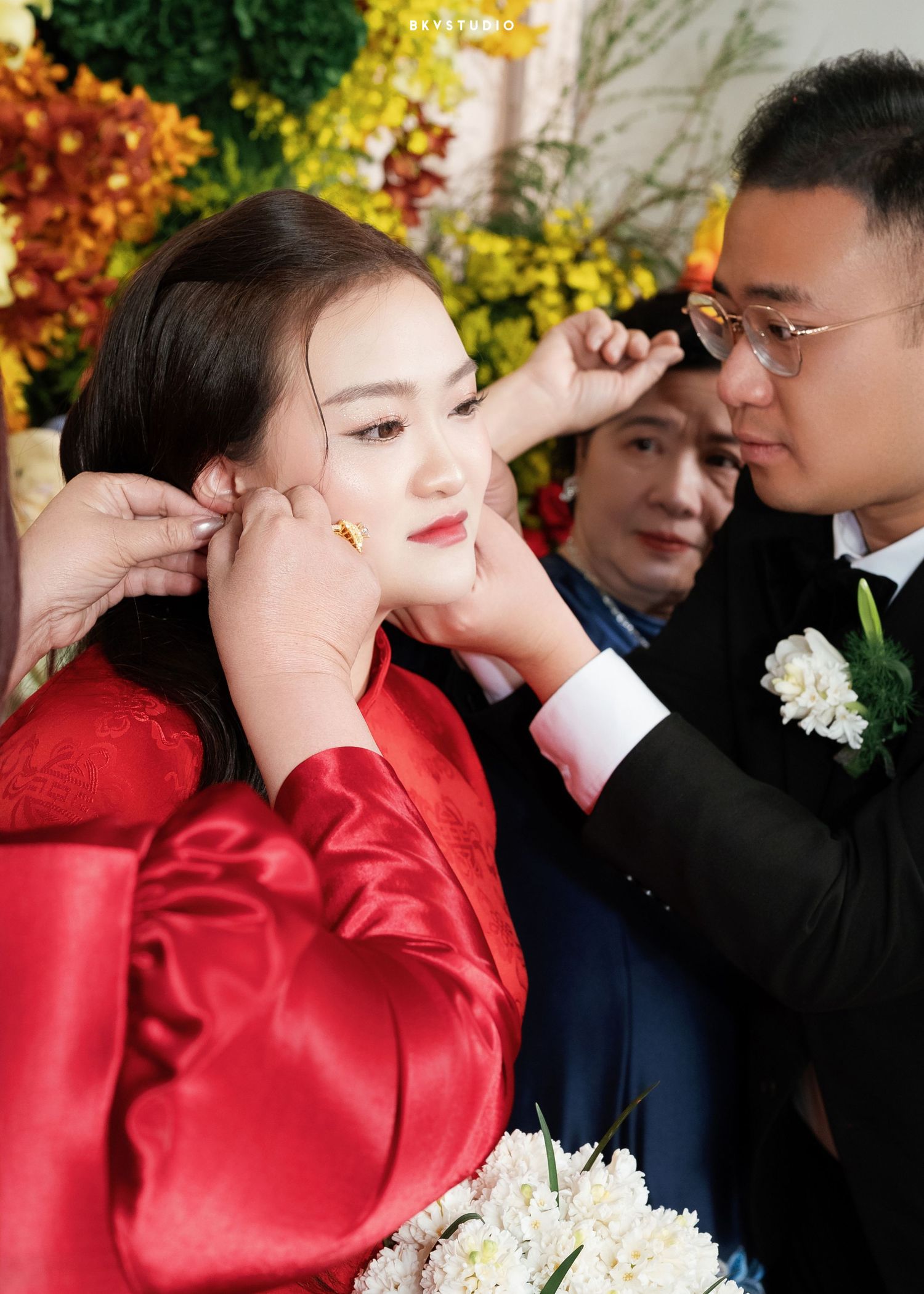 Groom placing gold earrings on bride in red traditional dress during Asian wedding ceremony with flowers in background.